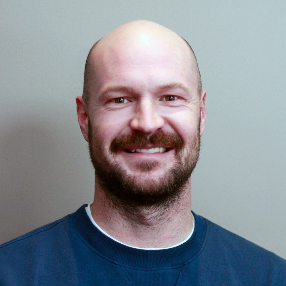 This image shows a smiling person with a bald head, a beard, wearing a navy blue shirt, in front of a grey background.