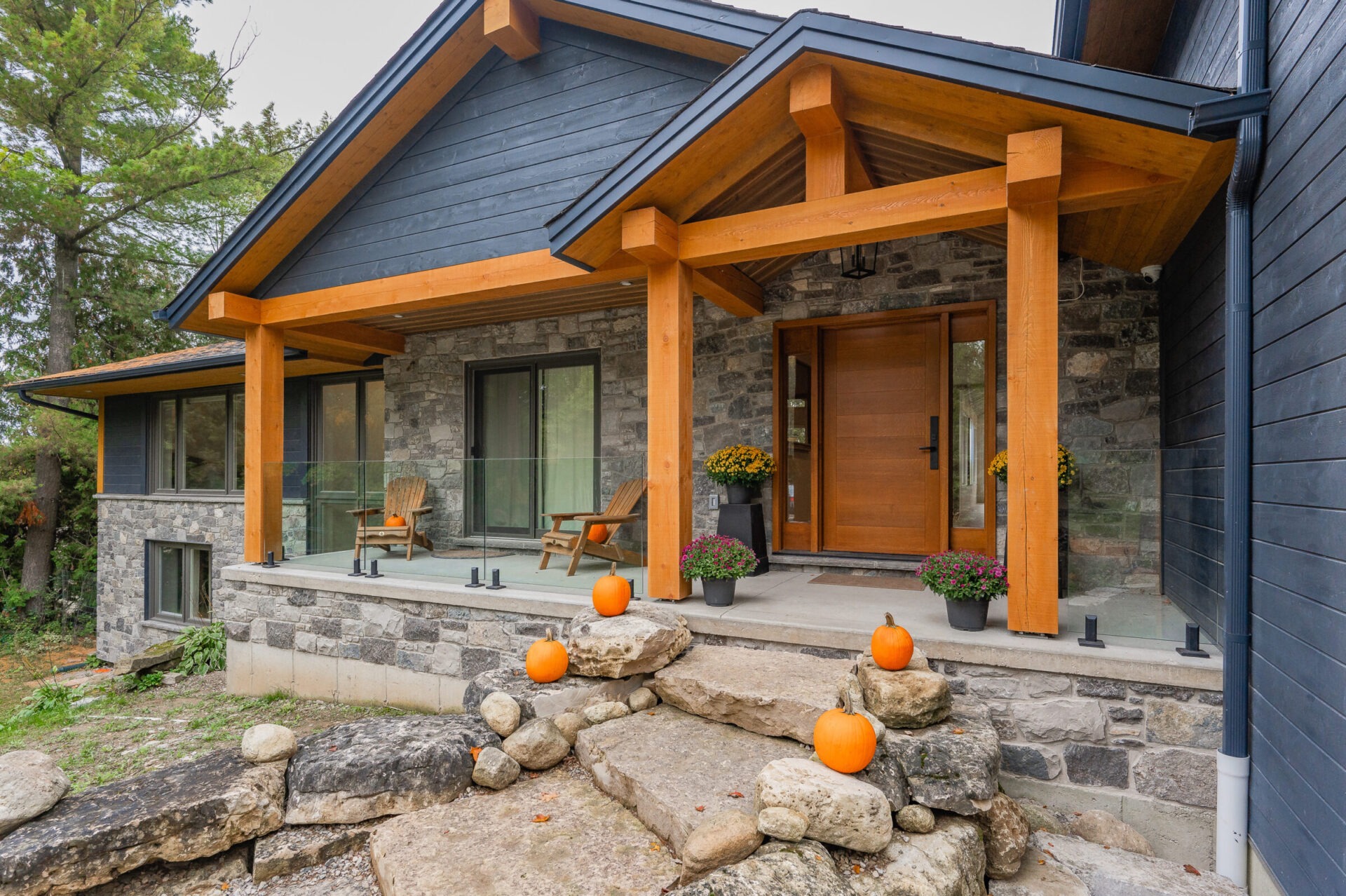 A modern house with a blue shingle roof, stone walls, wooden porch supports, a wood door, and seasonal decorations like pumpkins and flowers.