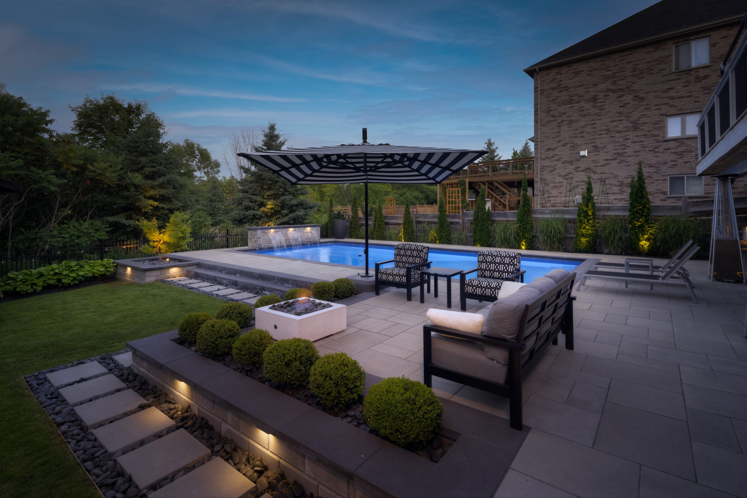 An elegant outdoor pool area at dusk, featuring stylish furniture, a fireplace, landscaping, and a modern house, creating a luxurious backyard atmosphere.
