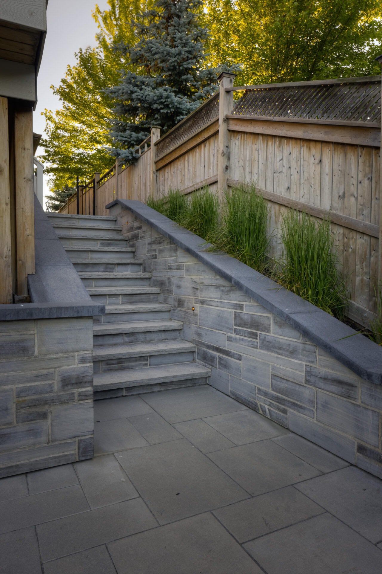 A stone stairway flanked by grass planters leads up between a wooden fence and a building, beneath trees with autumn foliage.