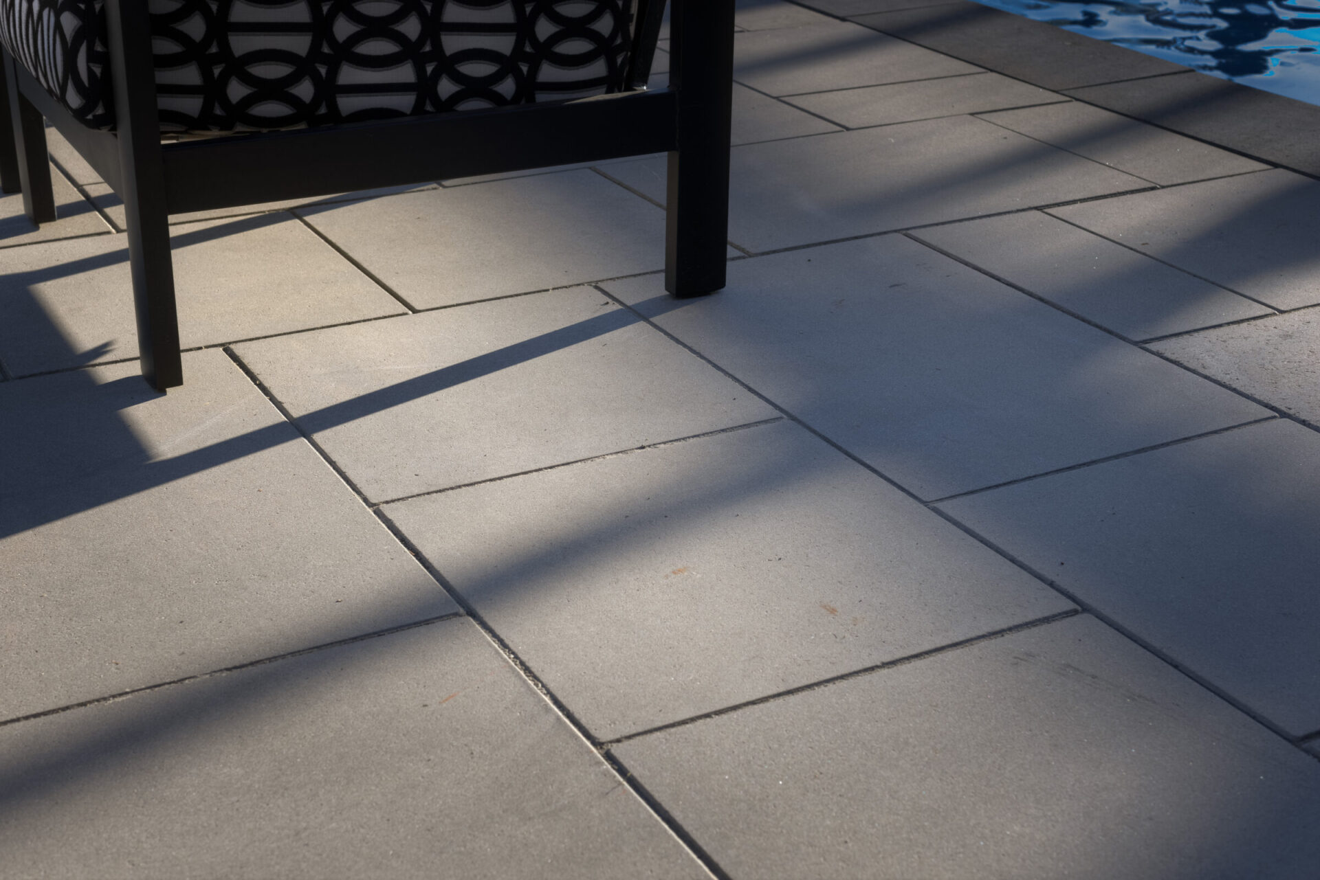 This image shows the floor of a patio with the shadow of a chair and part of a pool visible. The sunlight casts contrasting shadows on the tiles.