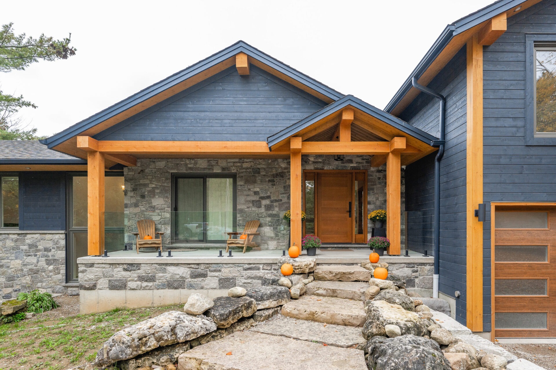 This image shows a modern house with dark blue siding, wooden accents, stone foundation, a porch with chairs, fall decorations, and a stone pathway.