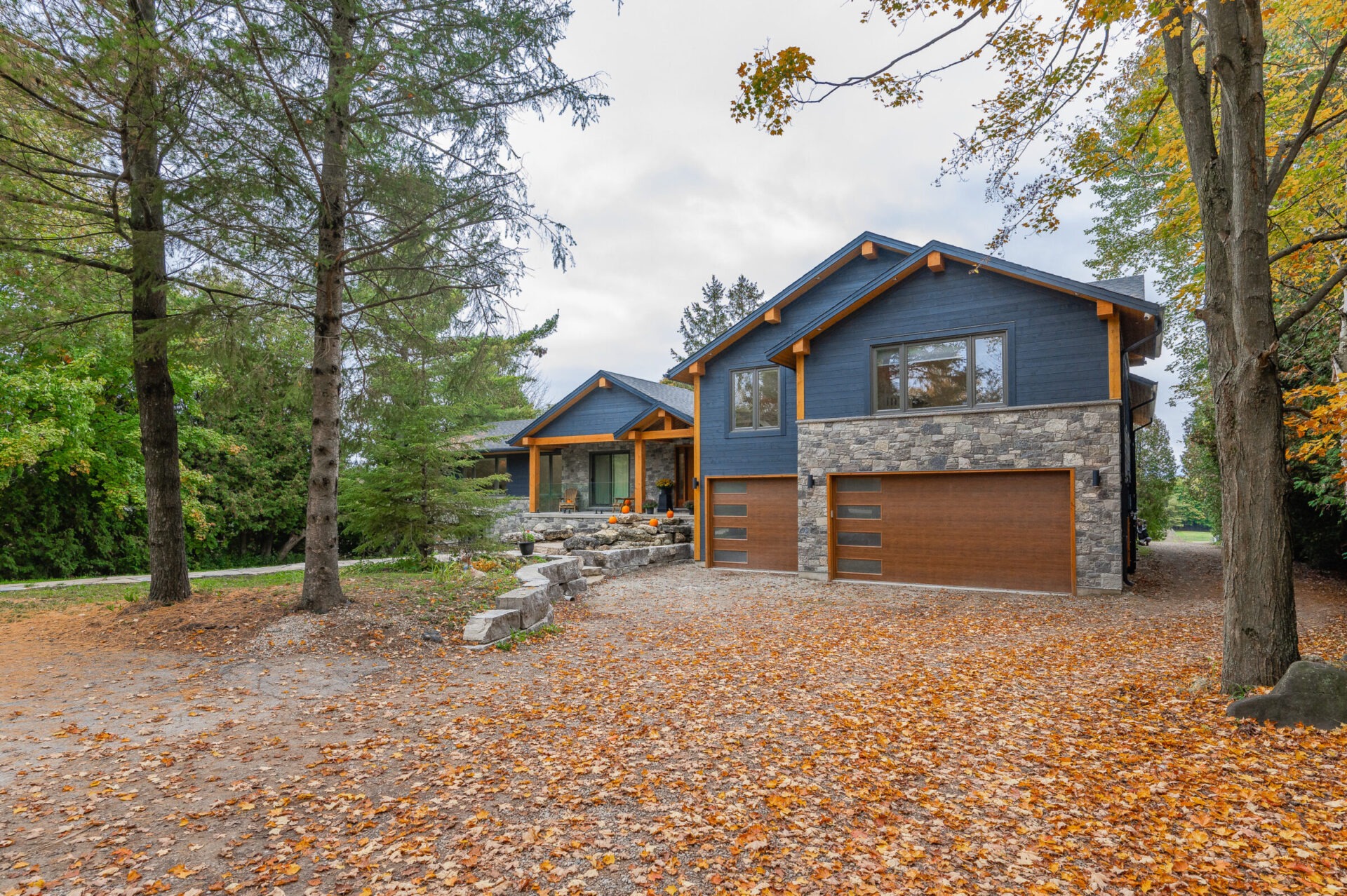 Modern two-story house with dark blue and stone facade, garage, surrounded by trees, with a driveway covered in fallen orange autumn leaves.