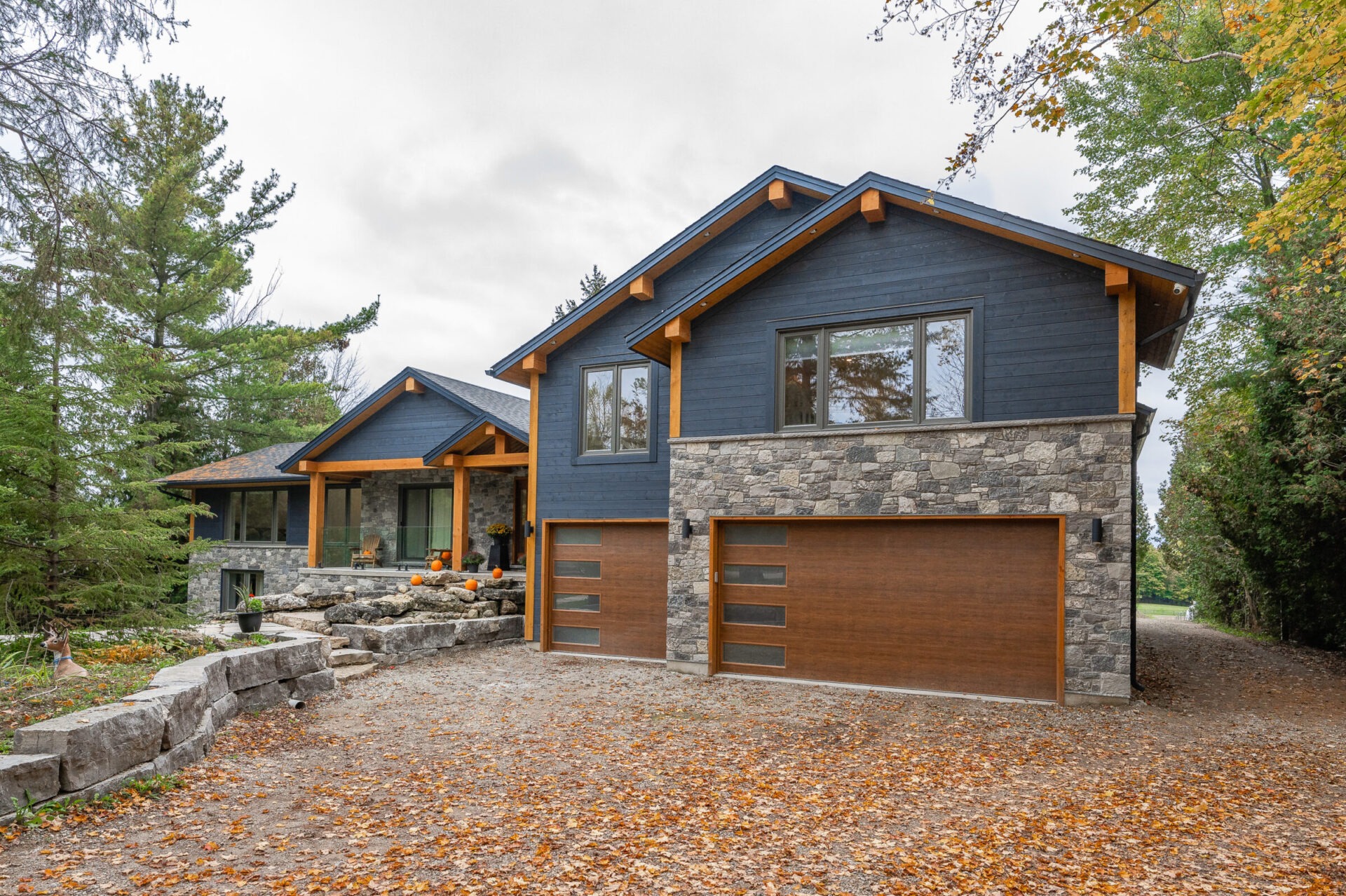 Modern house with dark blue siding, wooden accents, and stone foundation set in a wooded area during autumn with fallen leaves and seasonal decorations.