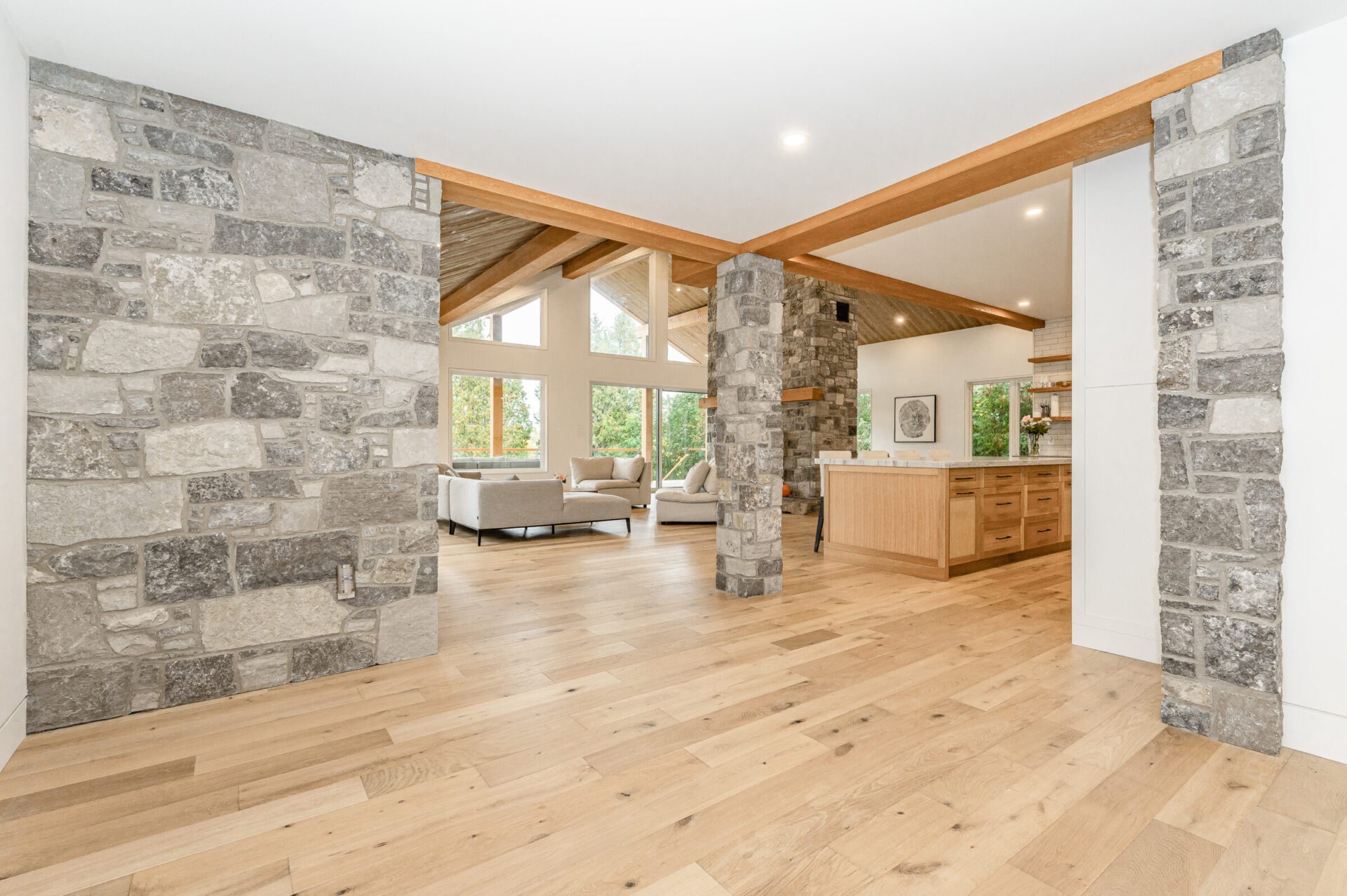 Modern spacious room with natural light, wooden floors and ceiling beams, stone columns, plush seating, and an open-plan kitchen in the background.