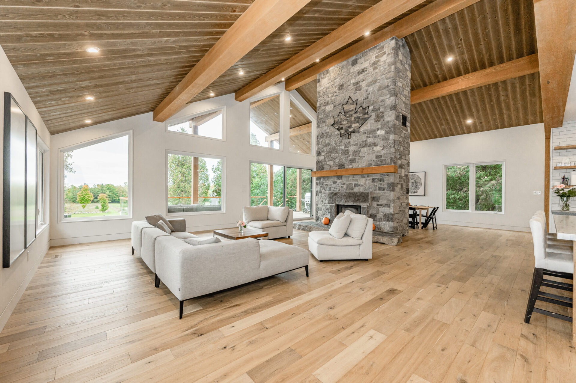 A spacious, modern living room with high ceilings, wooden beams, large windows, a stone fireplace, and contemporary furniture. Bright, natural light fills the space.