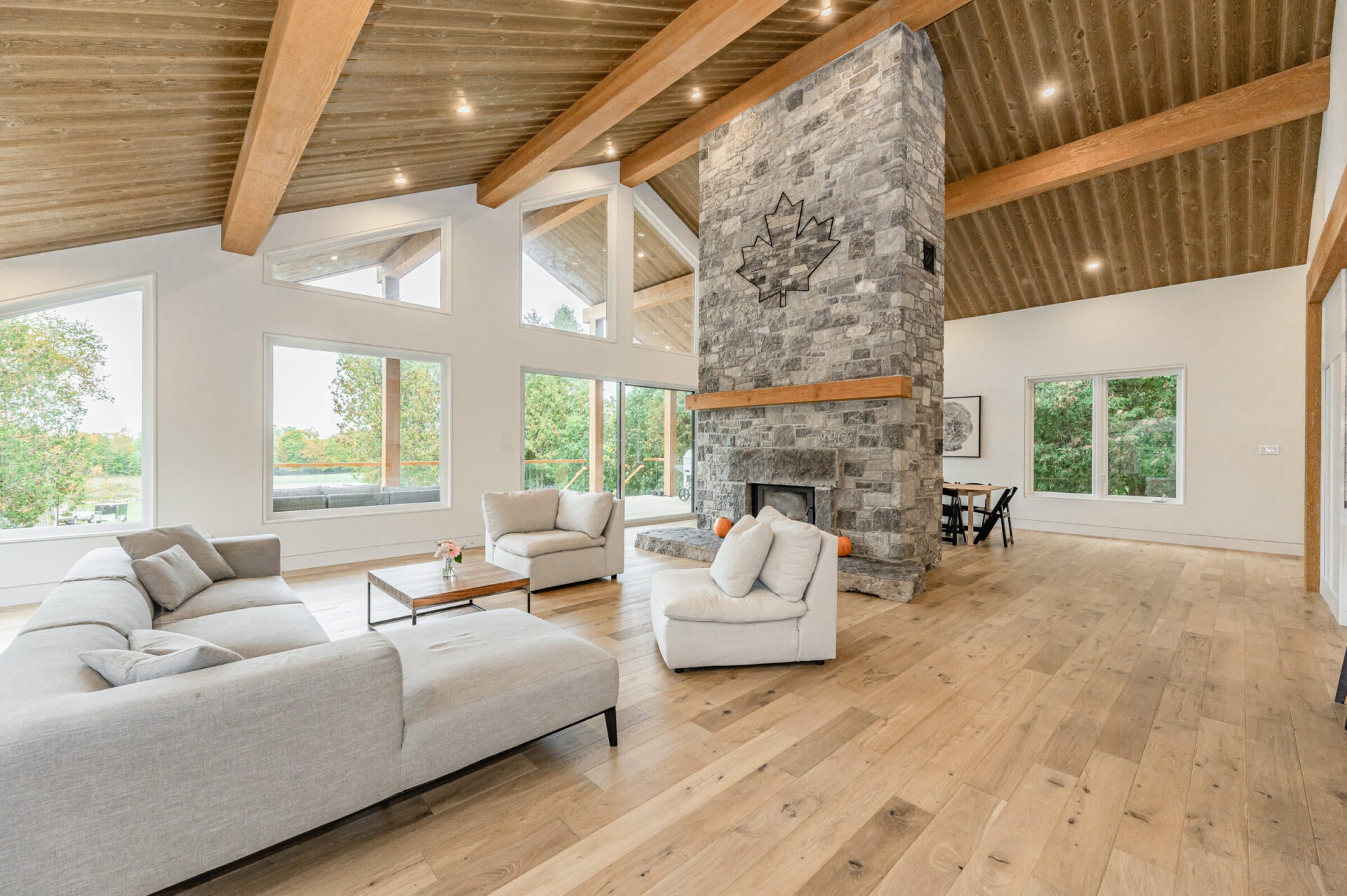 A spacious living room with high ceilings, exposed wooden beams, large windows, a stone fireplace, and minimalist furniture. It looks modern and airy.