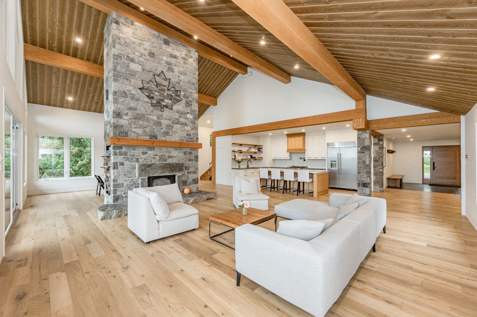 A spacious living room with a stone fireplace, exposed wooden beams, hardwood floors, white sofas, and an open-plan kitchen in the background.