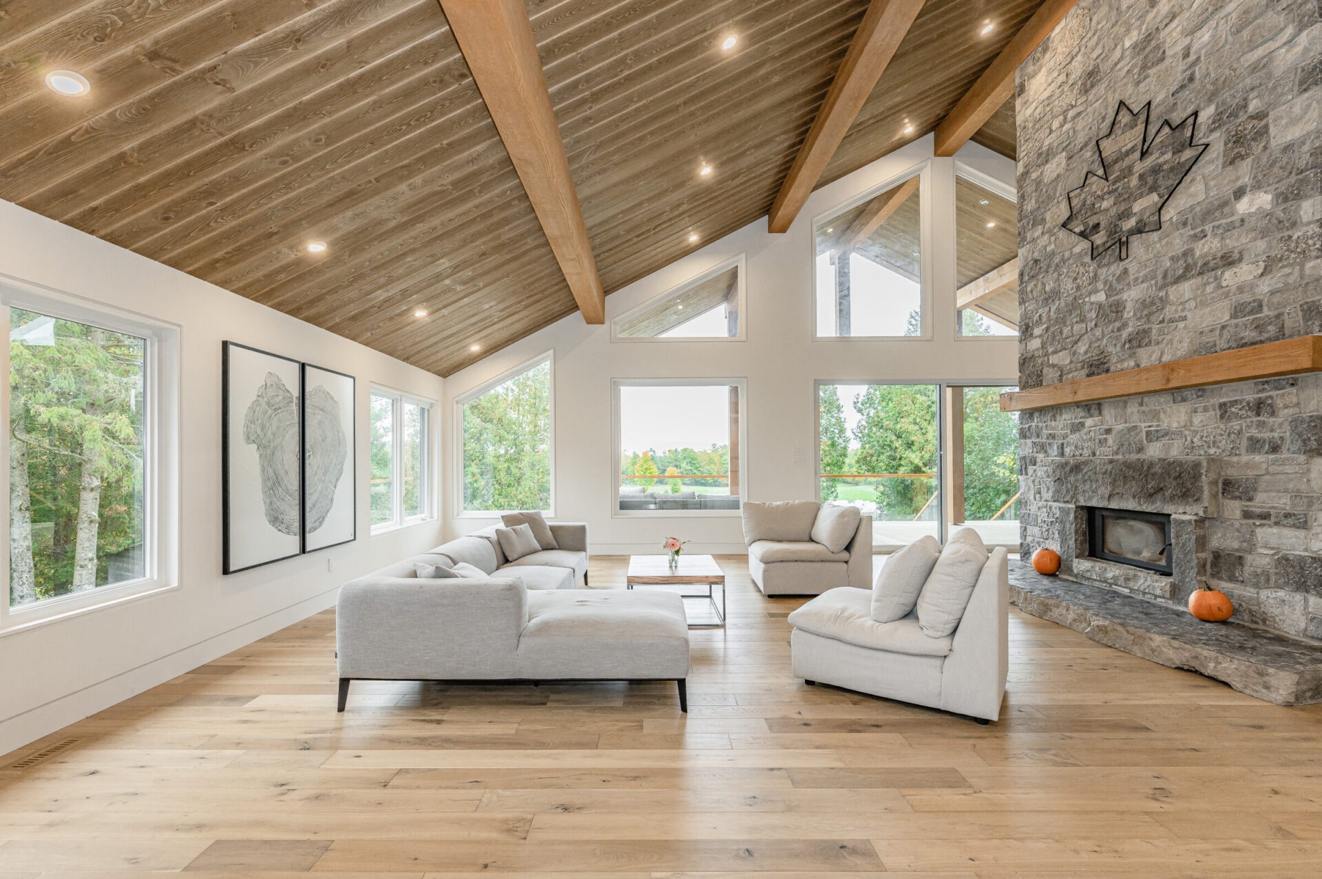 Spacious living room with high ceilings, exposed wooden beams, stone fireplace, large windows, hardwood floors, and neutral-toned furniture. Bright and modern interior.