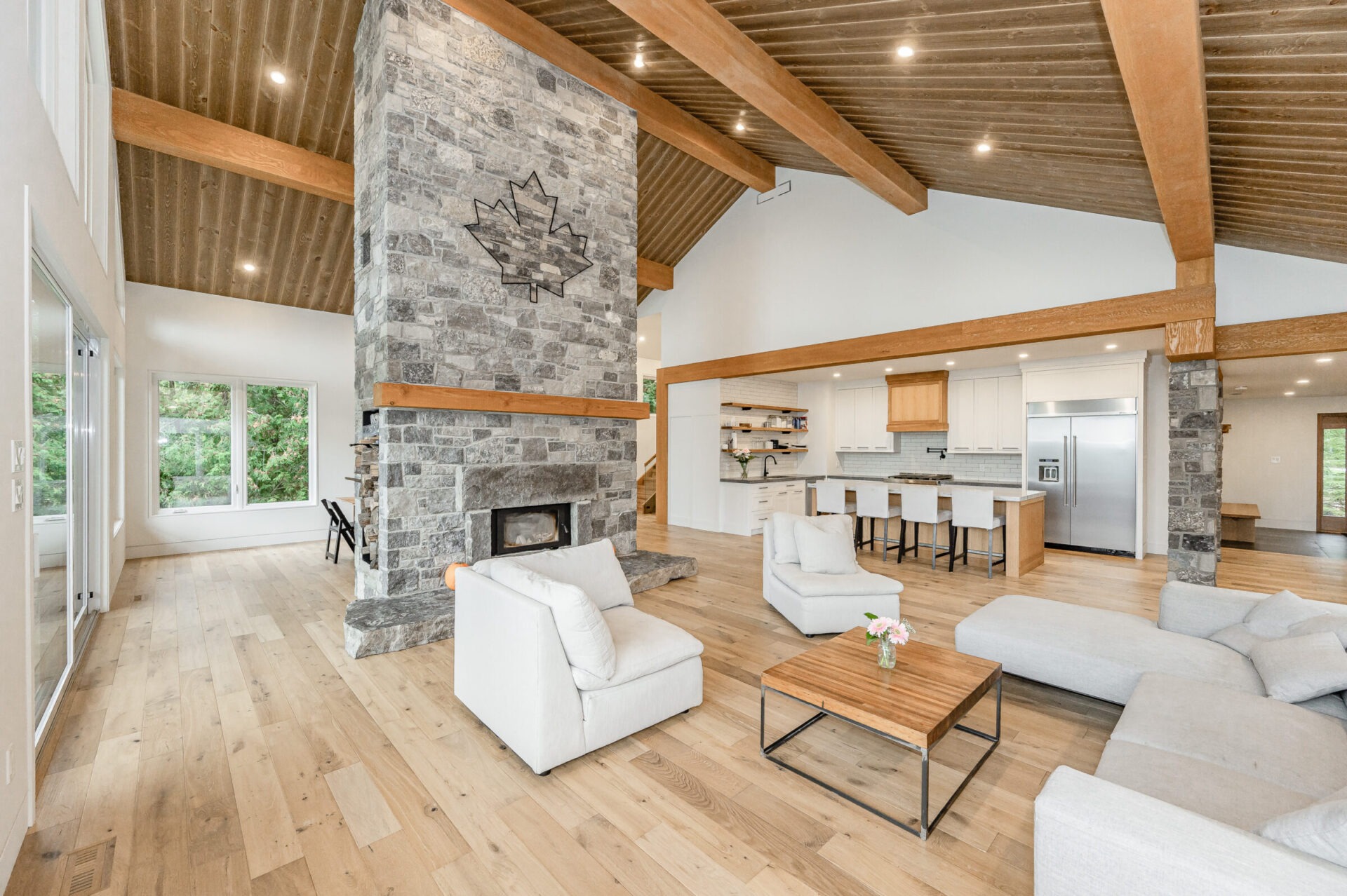 A spacious, modern living room with high ceilings, wooden beams, stone fireplace, open kitchen, hardwood floors, and contemporary furniture. Bright and airy ambience.