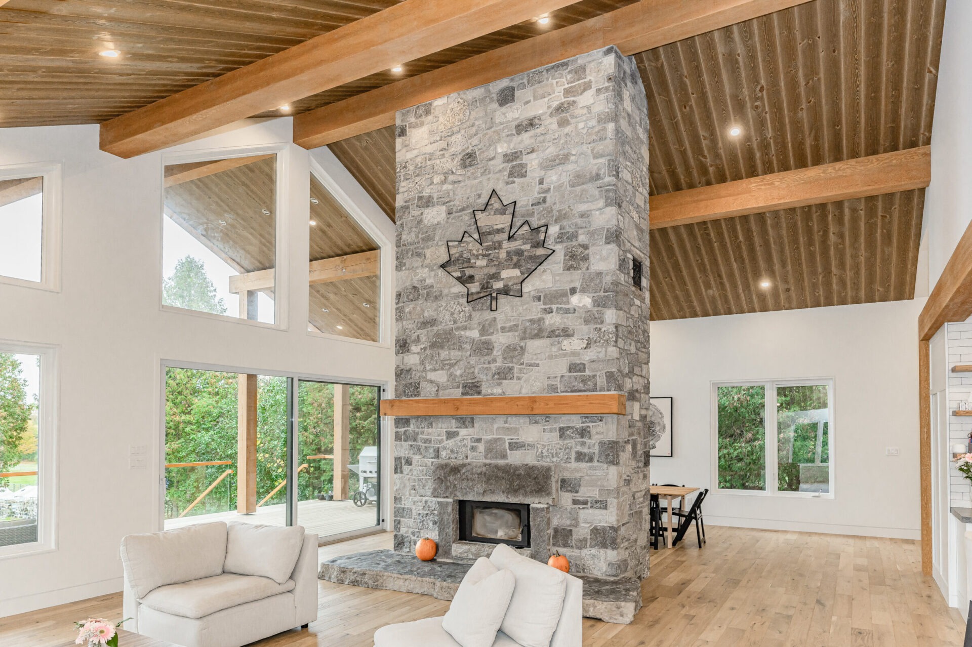 This is a bright, spacious living room with a large stone fireplace featuring a maple leaf design, wooden ceiling beams, and floor-to-ceiling windows.
