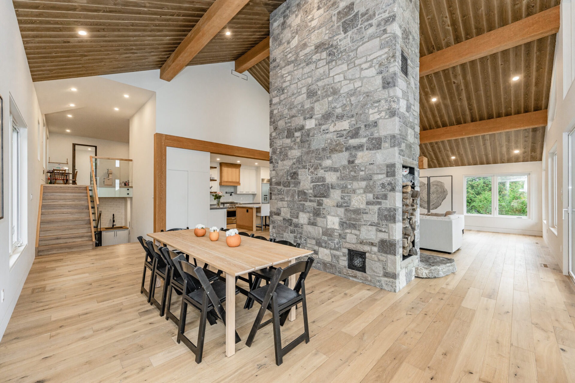 Modern interior with a wooden dining table, black chairs, stone fireplace, exposed beams, and an open plan layout leading to a kitchen and living area.