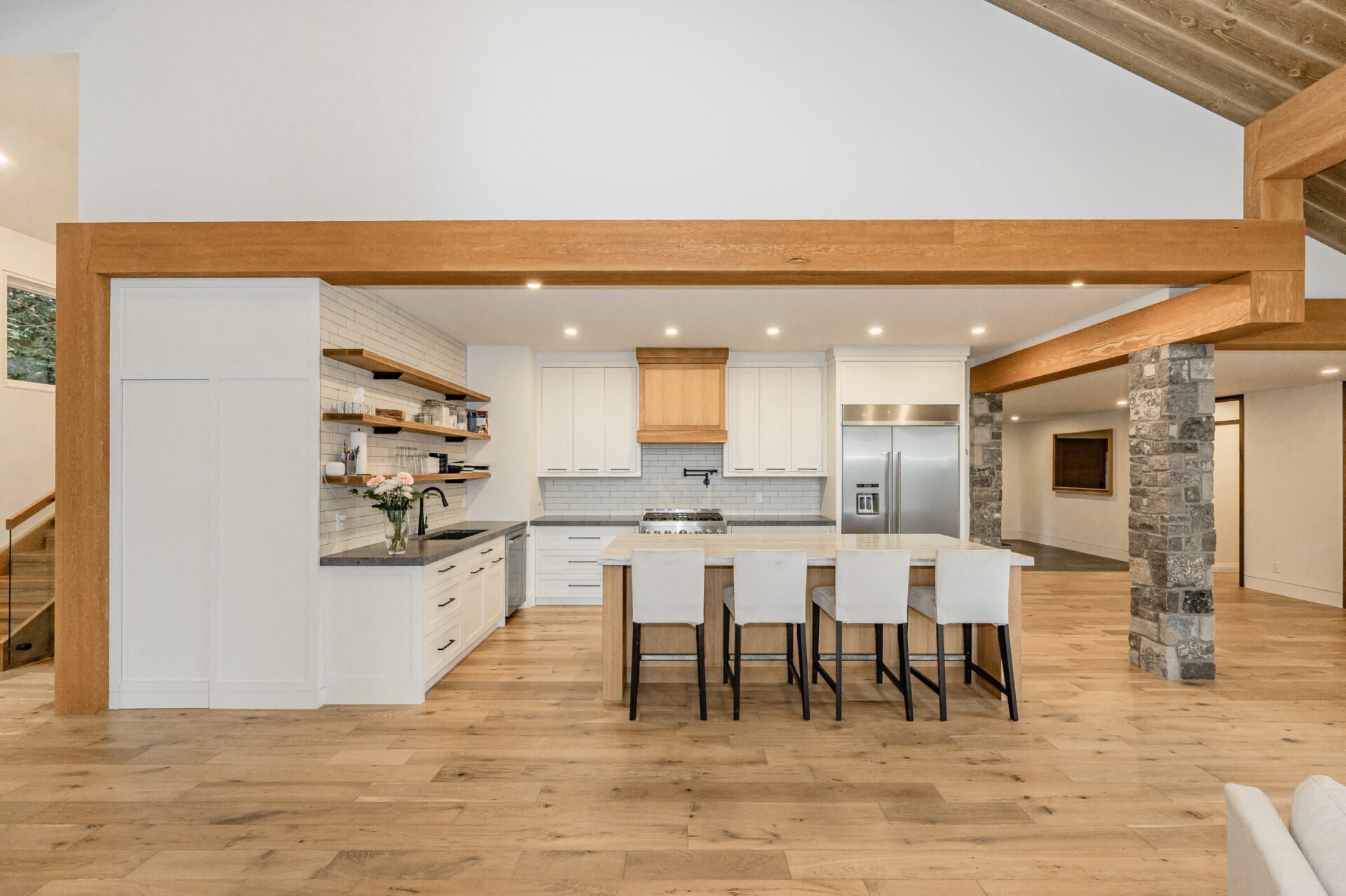 A spacious modern kitchen with white cabinets, stainless steel appliances, wooden beams, a stone column, and a central island with bar stools.
