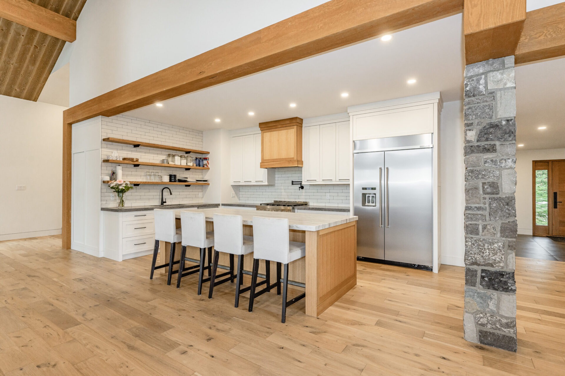A spacious kitchen with wooden floors, white cabinets, stainless steel appliances, a stone pillar, and a center island with seating. Natural light enhances the warm ambiance.