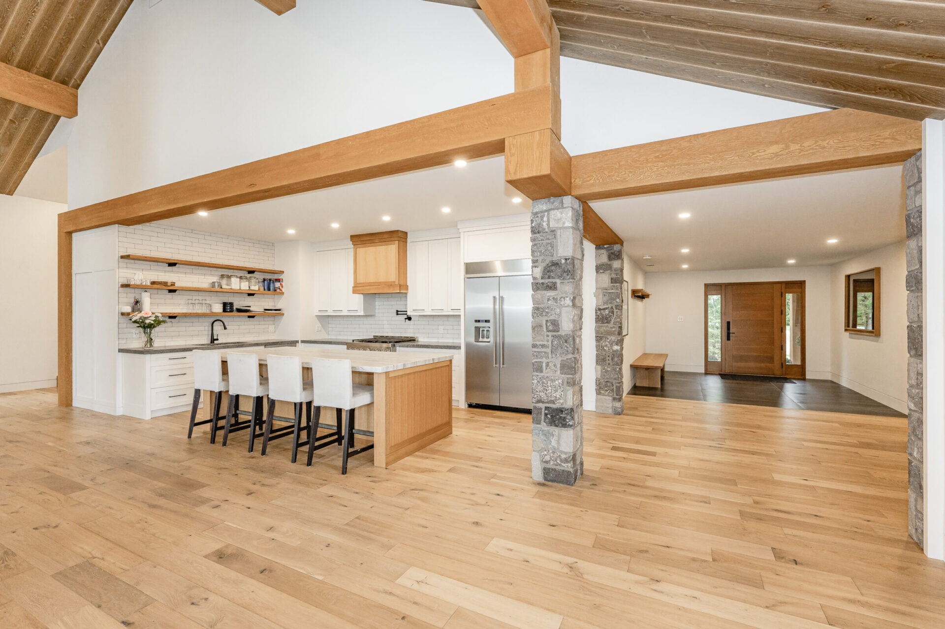 A spacious, modern kitchen with white countertops, wooden beams, stone pillars, bar stools, and stainless steel appliances. Hardwood flooring throughout with ample light.