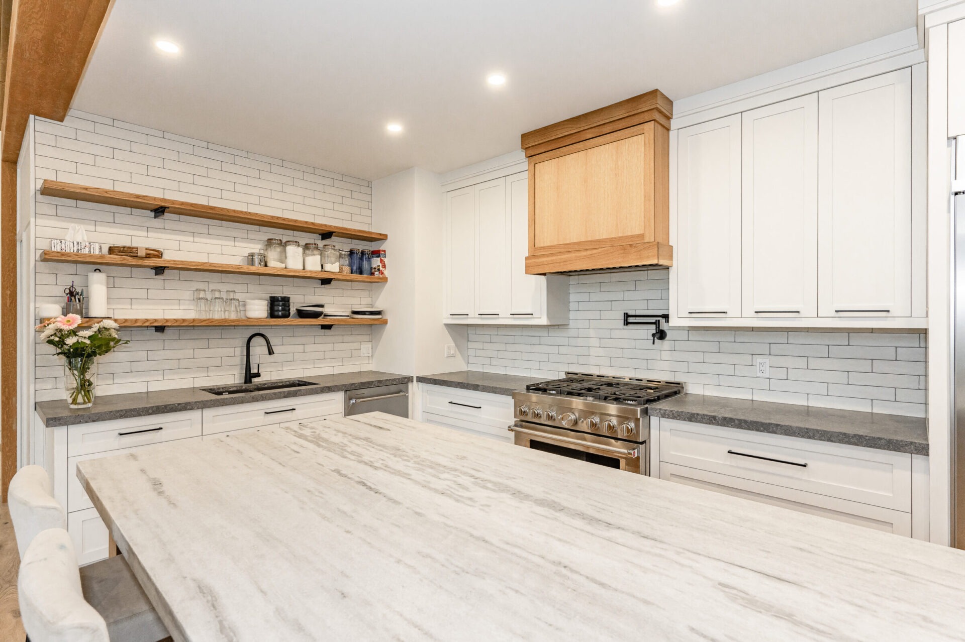 Modern kitchen interior with white cabinetry, subway tile backsplash, wooden shelves, stainless steel stove, and marble countertops with natural light.