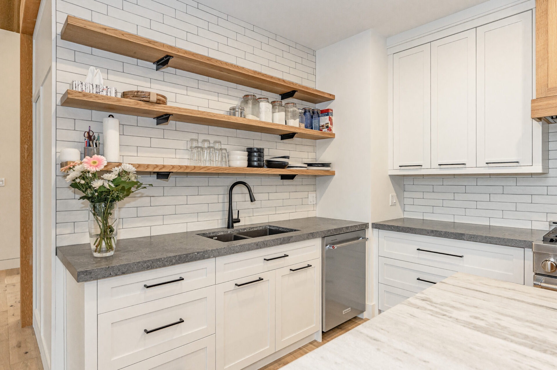 A modern kitchen with white cabinetry, gray countertops, subway tile backsplash, open wooden shelves, and stainless-steel appliances. A vase with flowers adds color.