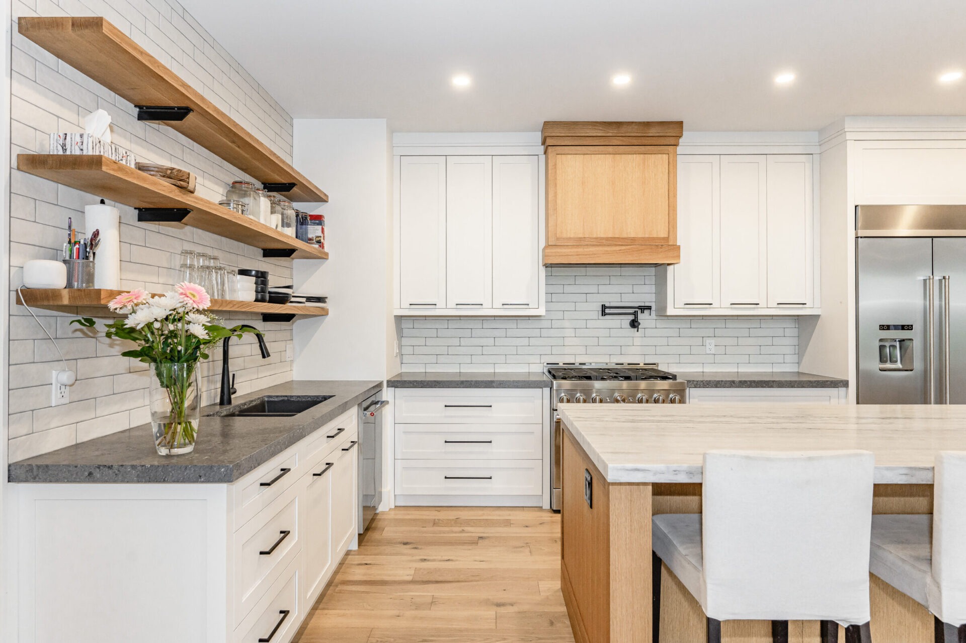 A modern kitchen with white cabinetry, stainless steel appliances, floating wooden shelves, a subway tile backsplash, and an island with seating.