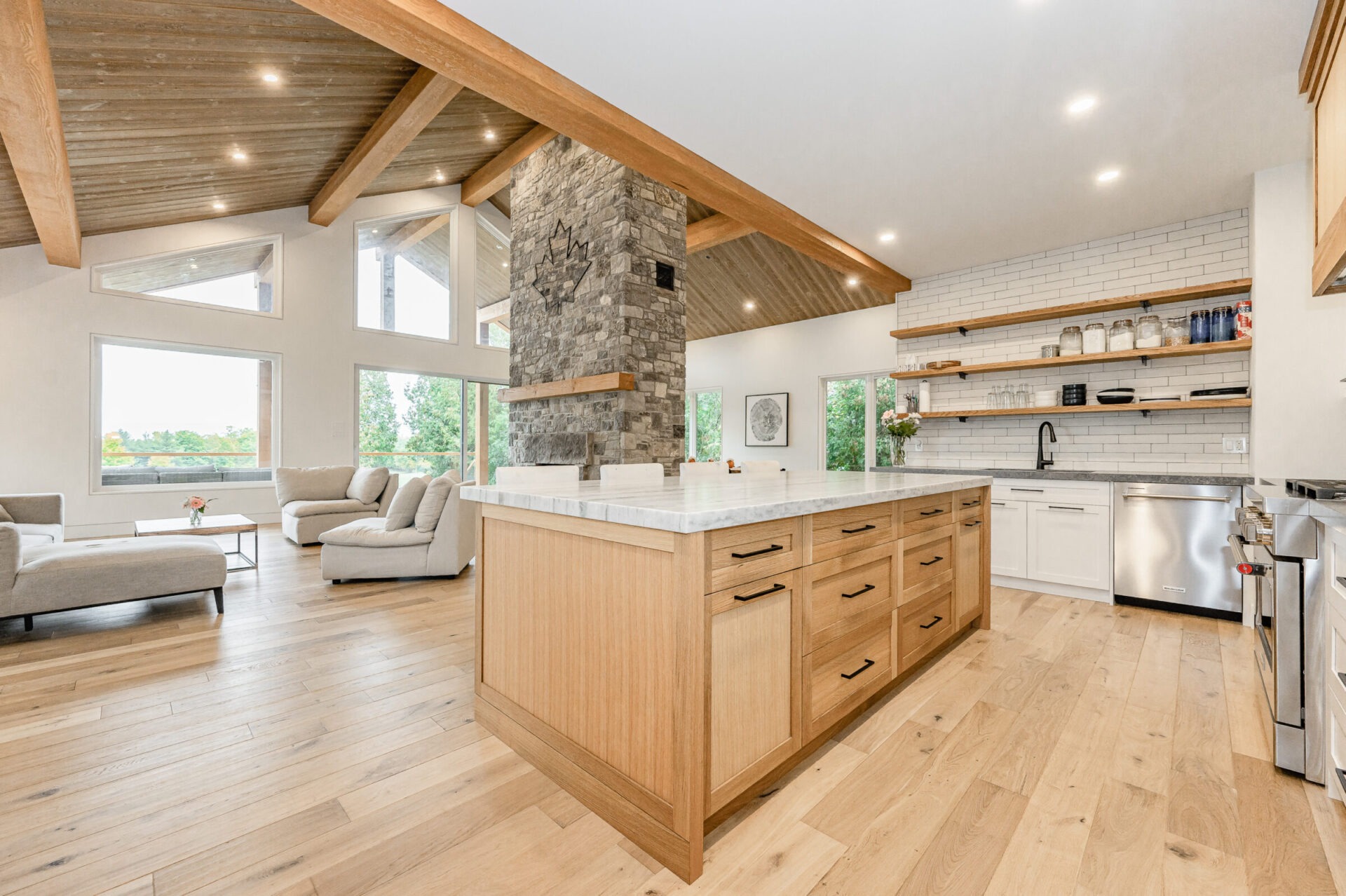 Modern open-plan kitchen with wooden cabinets, stone column, stainless steel appliances, white subway tile backsplash, and adjacent living area with sofa.