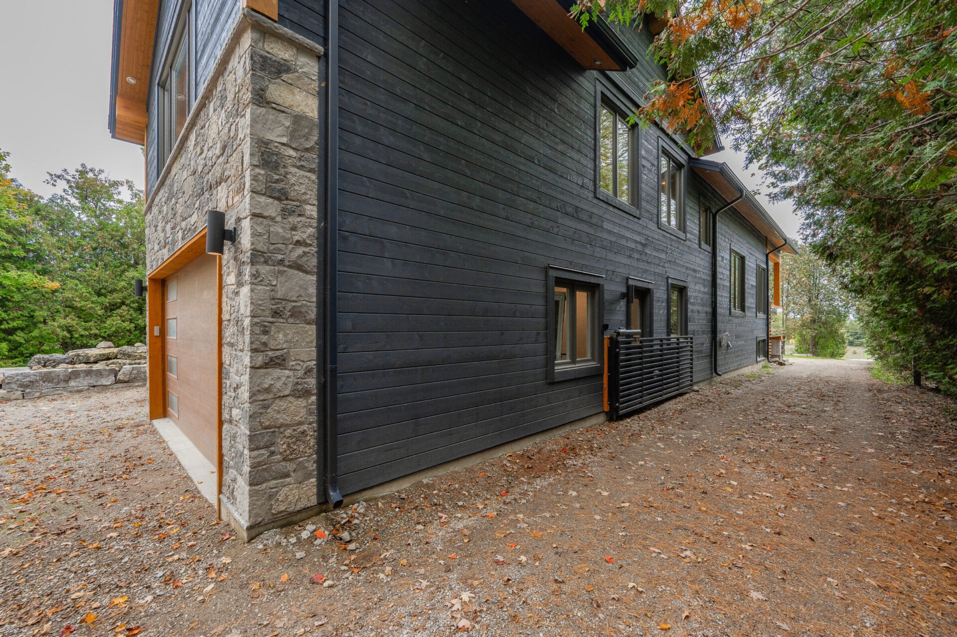 Modern two-story house with dark siding and stone accent wall, surrounded by trees with a gravel path leading to the entrance.