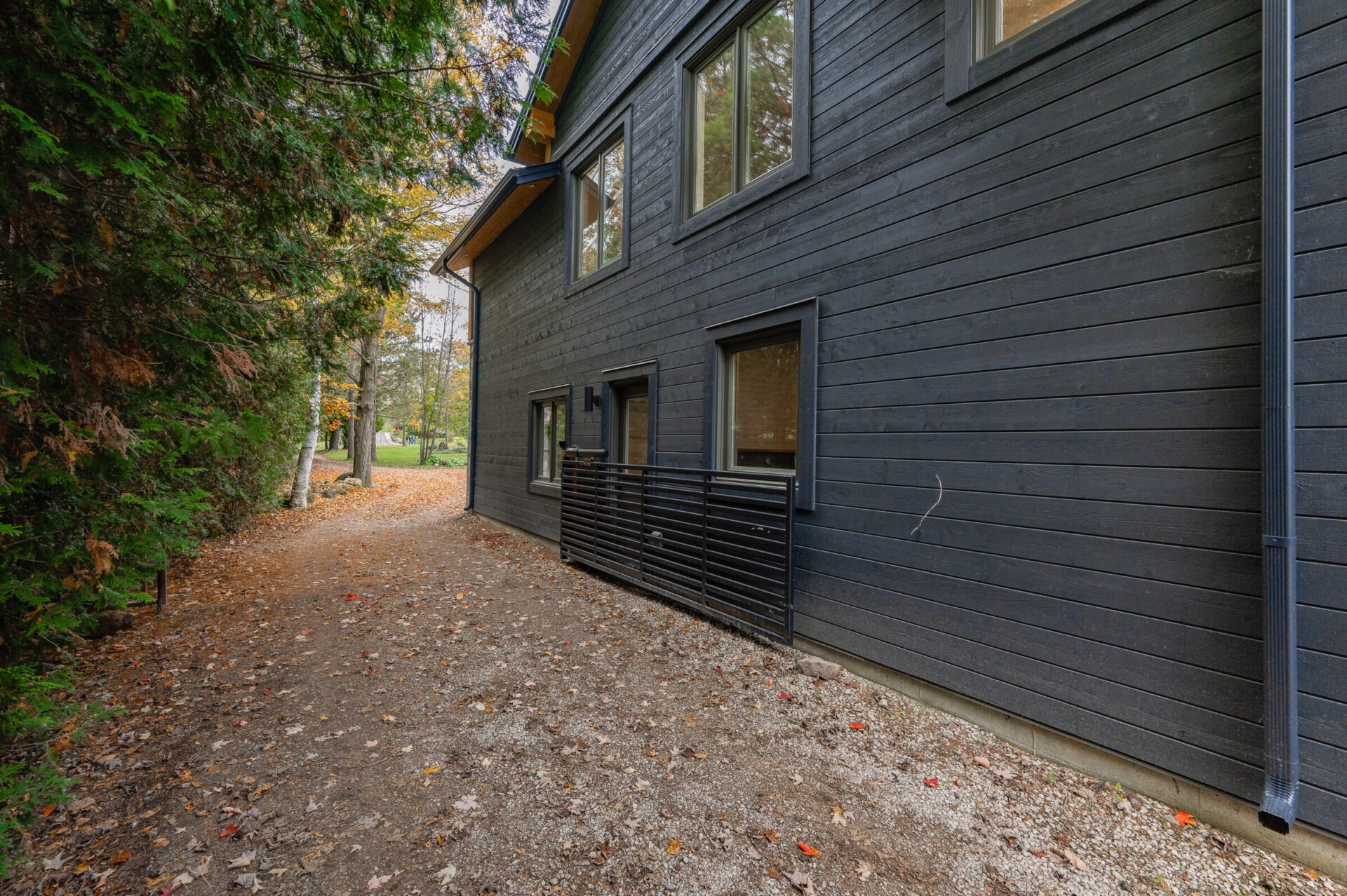 A modern two-story house with dark siding is shown next to a gravel path, surrounded by trees and scattered autumn leaves.