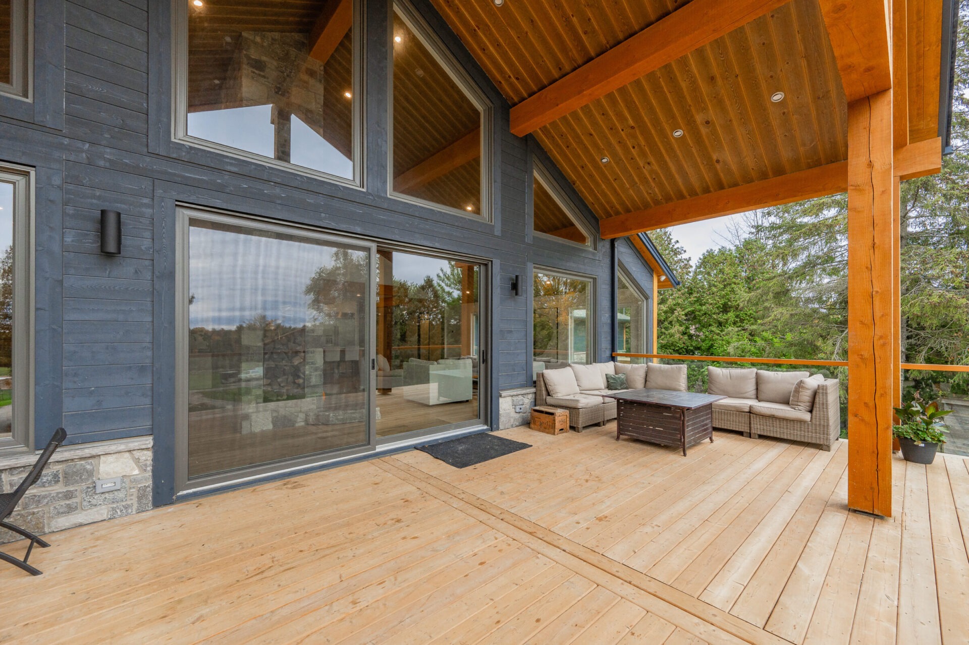 This is a spacious wooden deck of a modern house with large glass windows, comfortable outdoor furniture, and a view of greenery outside.