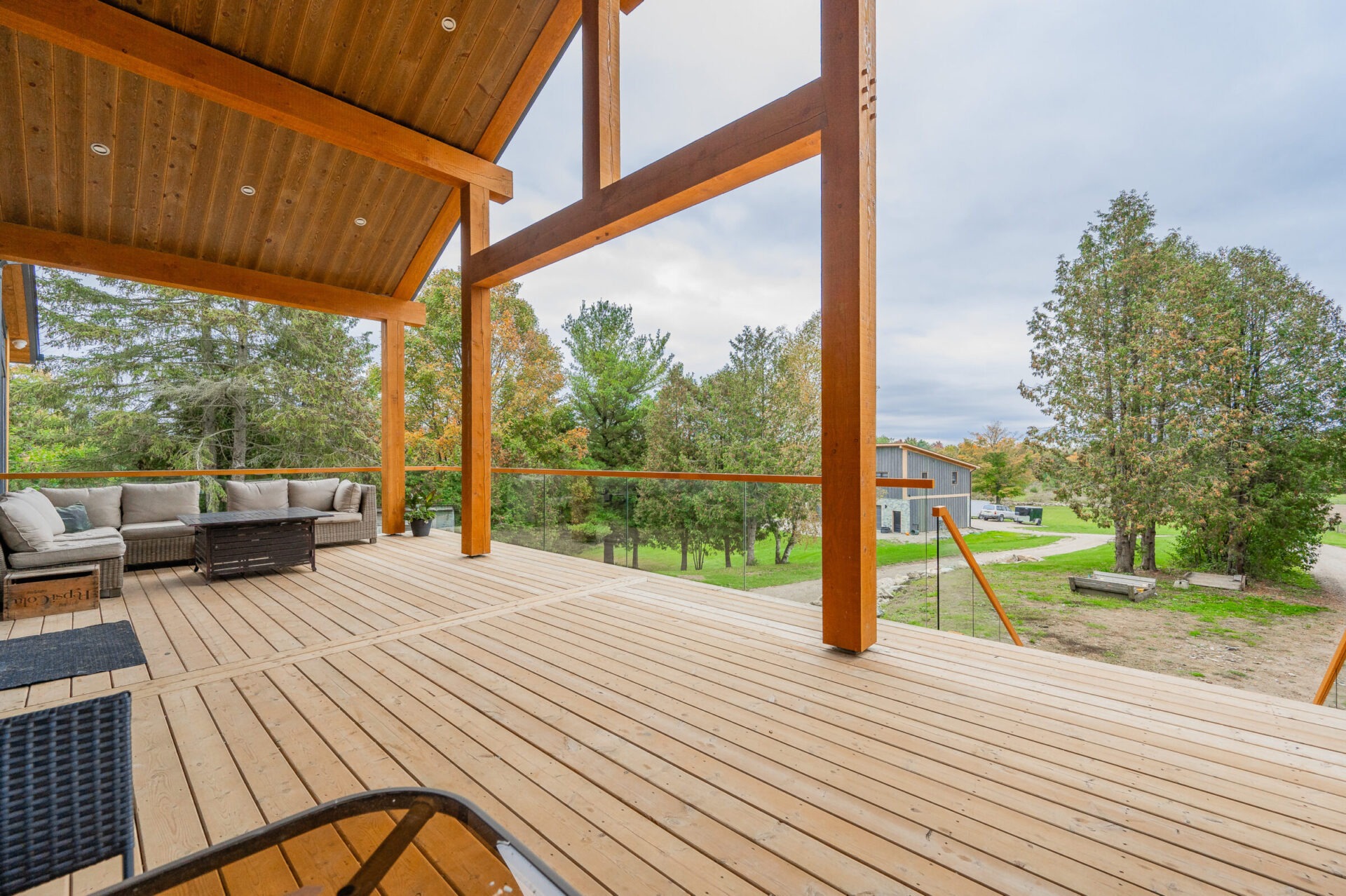 A spacious wooden deck with outdoor furniture overlooks a serene landscape with trees and a grassy area. The deck has a covered section with exposed beams.