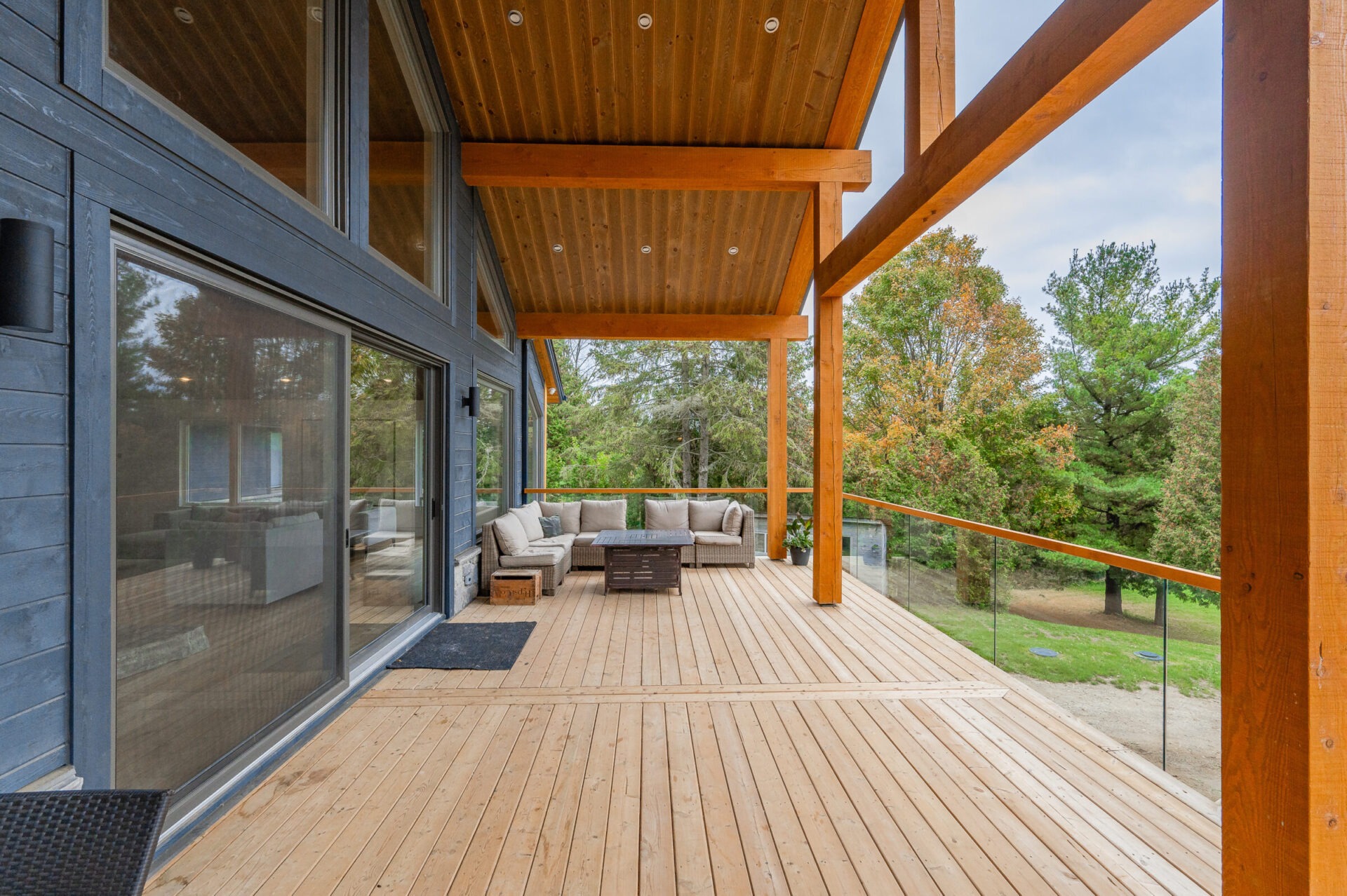 A spacious wooden deck with modern outdoor furniture overlooks a tranquil forest. The home features large glass doors and a wooden beam structure.