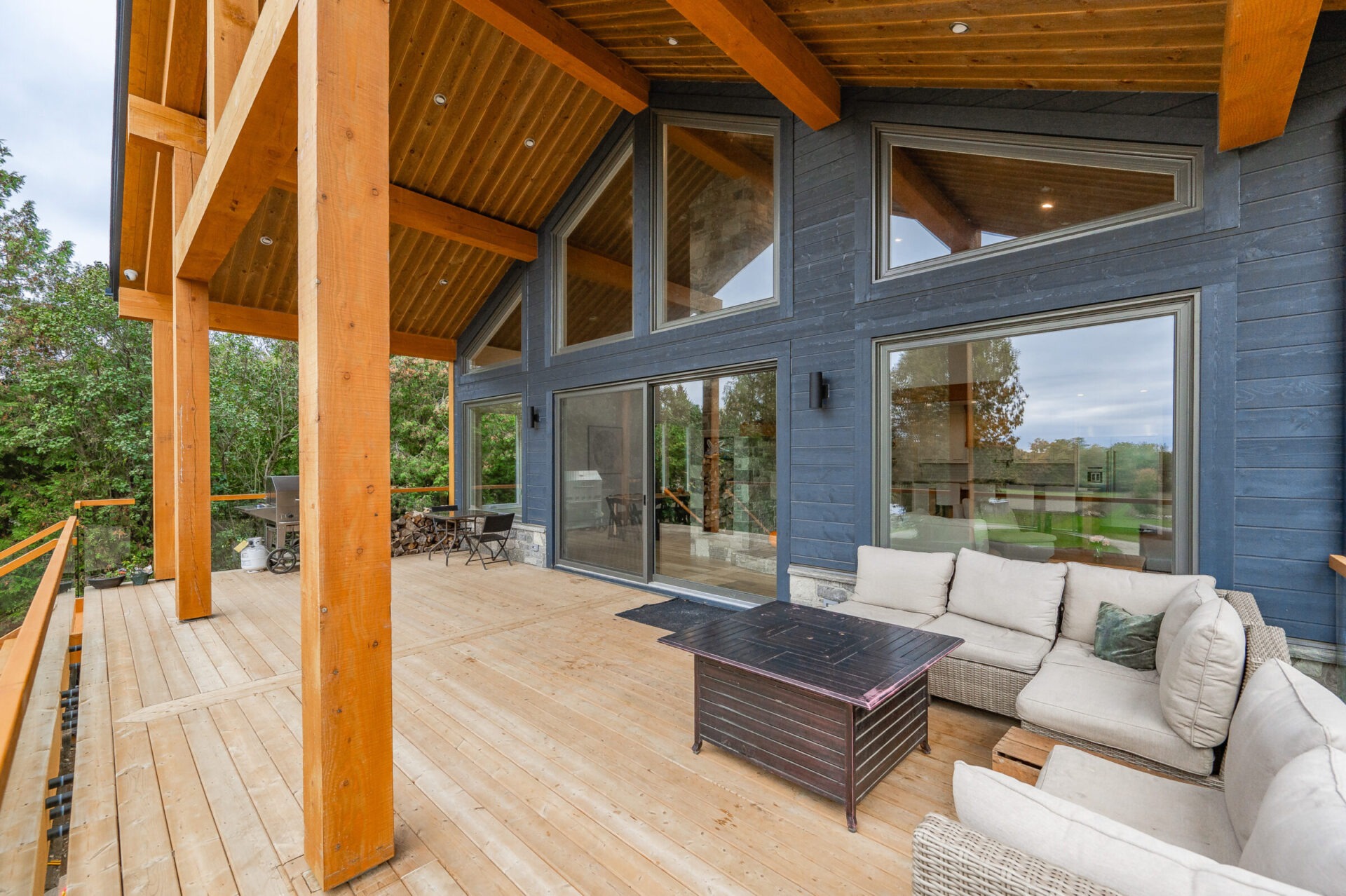 A spacious wooden deck features an outdoor seating area with cream cushions, a coffee table, and large windows on dark blue walls.