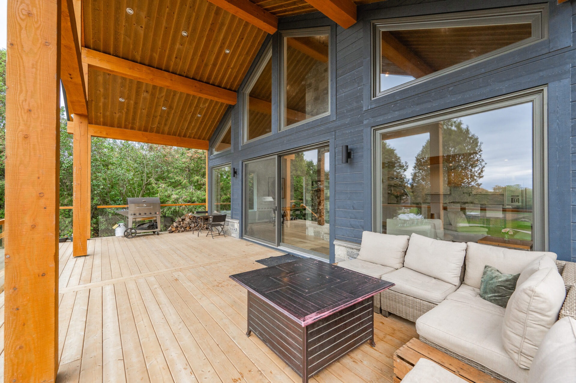 A cozy covered wooden deck features a rattan corner sofa, a grill, and overlooks a lush green yard with large windows reflecting the outdoors.