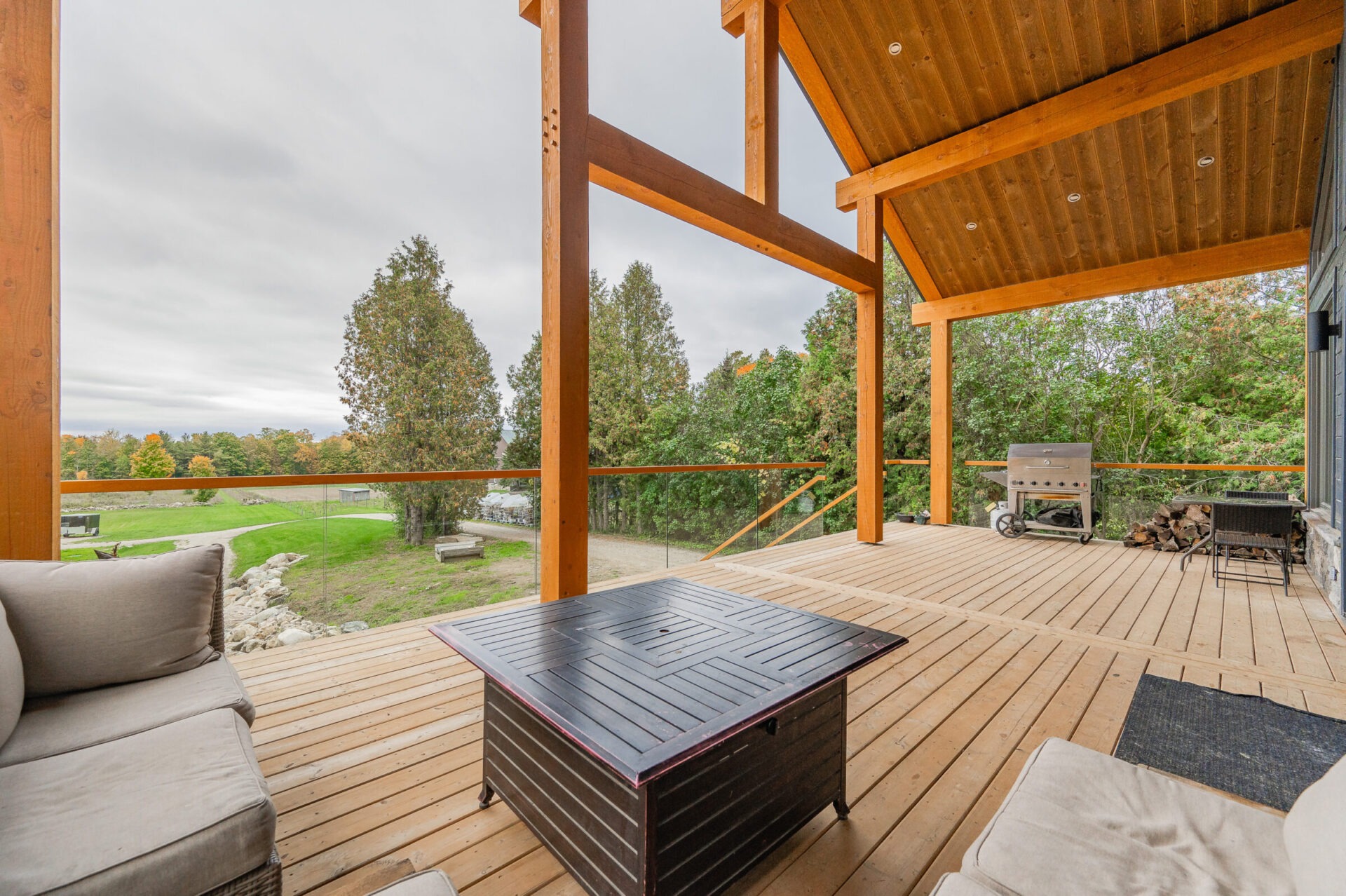 This image shows a spacious wooden deck with a comfortable seating area, a firepit table, and a tranquil view of a green landscape.