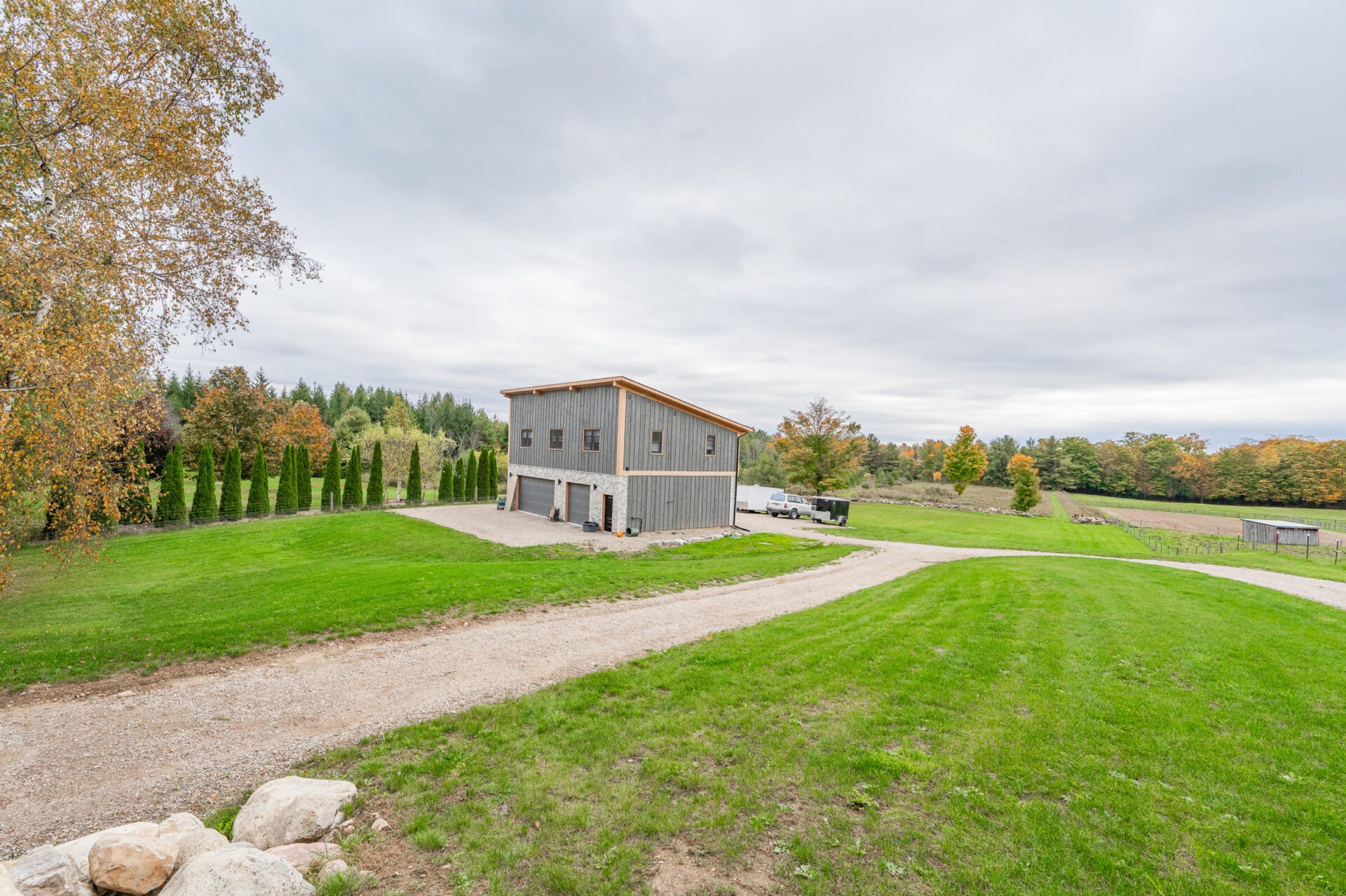 This is a rural scene with a large, two-story wooden building, a well-manicured lawn, gravel driveway, and neatly aligned trees under a cloudy sky.