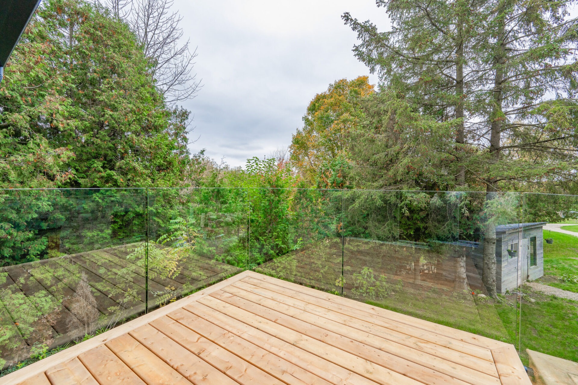 A spacious wooden deck with transparent glass balustrades overlooks a lush backyard with trees and a small garden shed, under a cloudy sky.