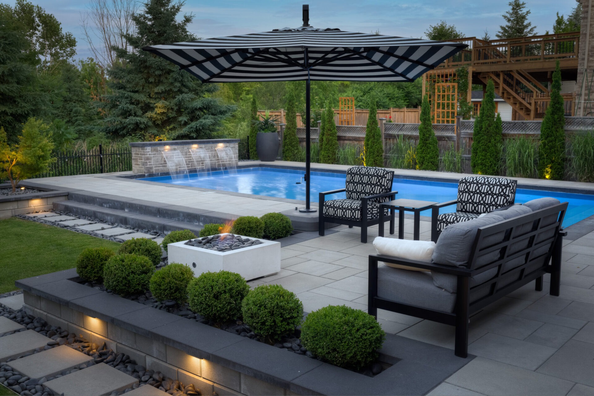 A luxurious backyard with a swimming pool, water features, stylish outdoor furniture, a striped umbrella, fire pit, manicured bushes, and a wooden deck.