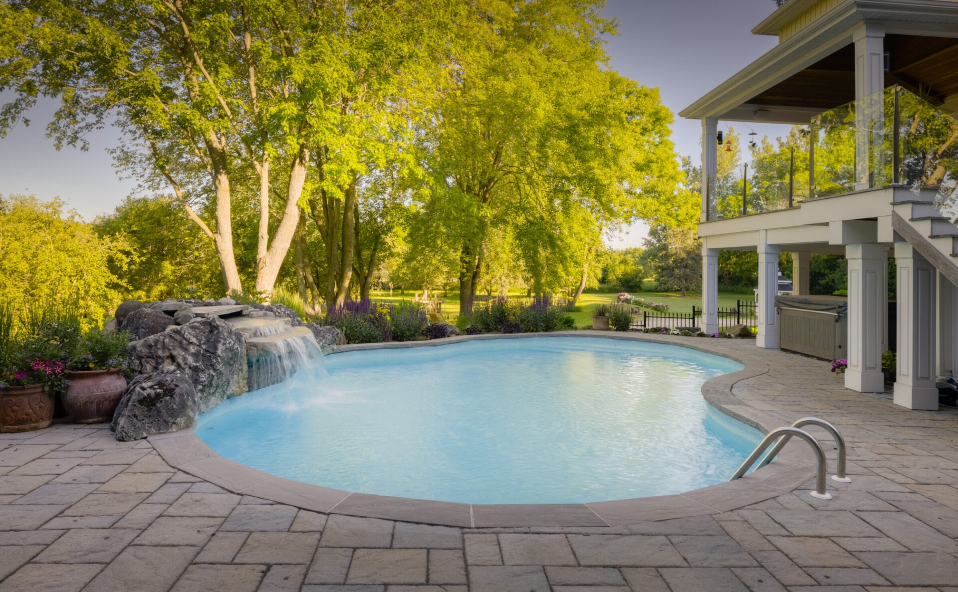 An idyllic backyard with a curved swimming pool, artificial waterfall, paver patio, and greenery under a clear sky, adjacent to a two-story house with porches.