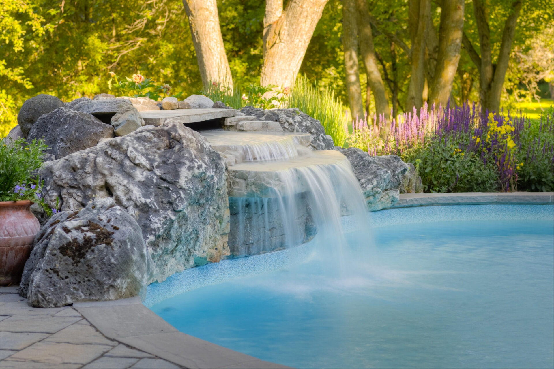 An artificial waterfall flows into a serene blue pool surrounded by lush greenery, purple flowers, large rocks, and a tranquil natural setting.