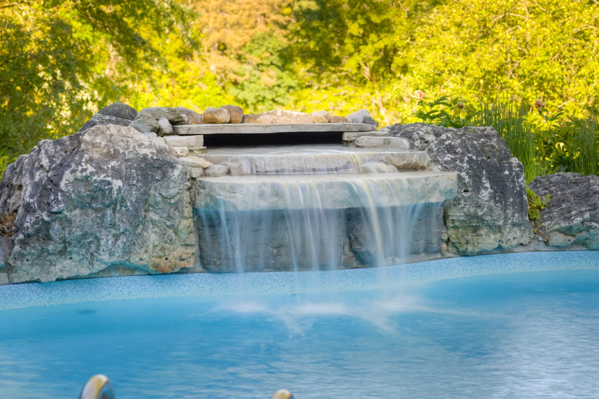 A manmade waterfall with clear cascading water over rocks into a tranquil blue pool, surrounded by lush green foliage in a serene setting.