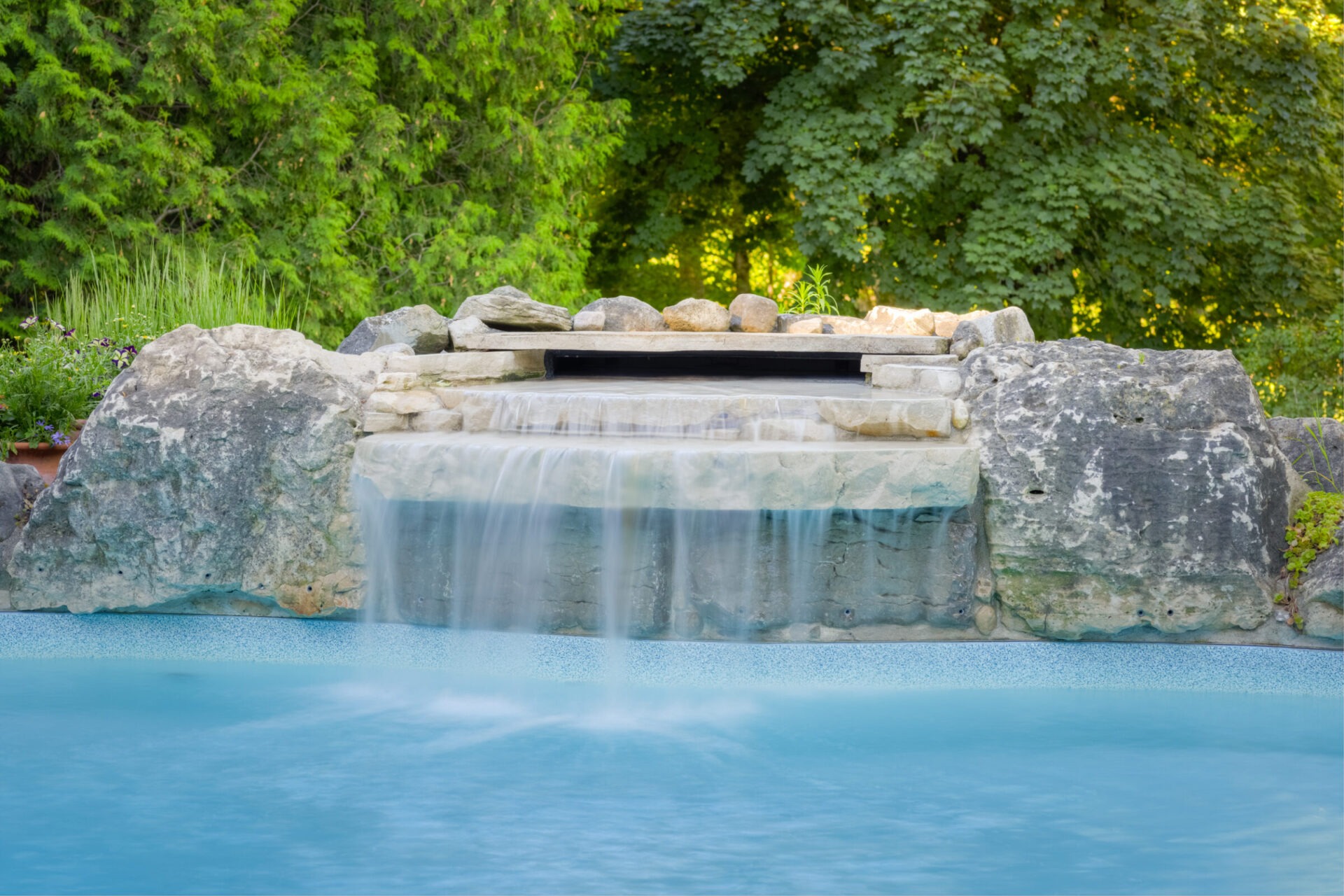 This image shows a serene pool with a built-in waterfall surrounded by natural rocks and lush green trees, evoking a tranquil, landscaped garden atmosphere.