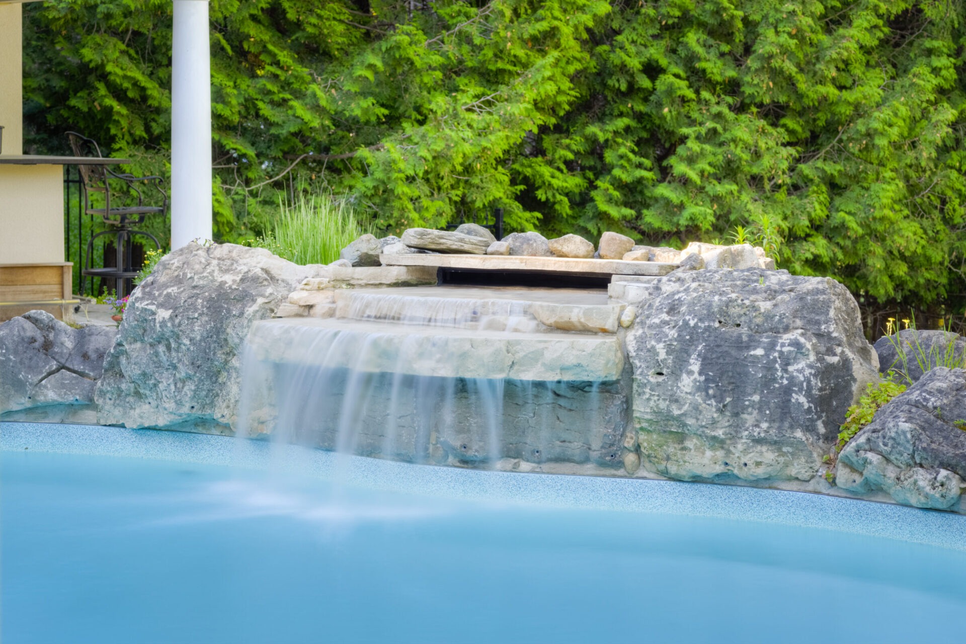 A serene artificial waterfall crafted from large stones pours into a clear blue pool, surrounded by lush greenery and a partial building view.