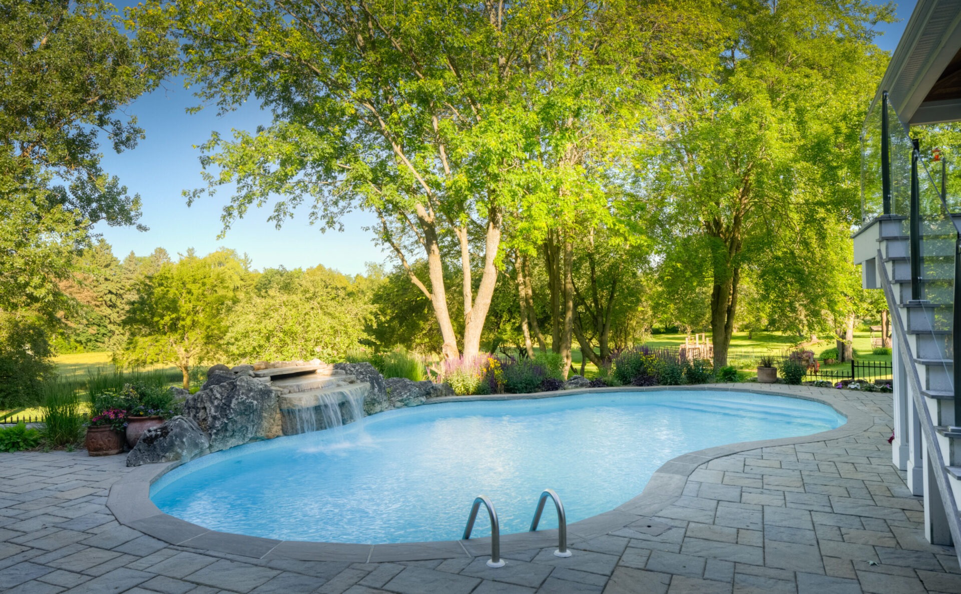 This image shows a tranquil backyard with a curvy swimming pool, stone waterfall feature, paved patio, lush greenery, and clear blue skies.