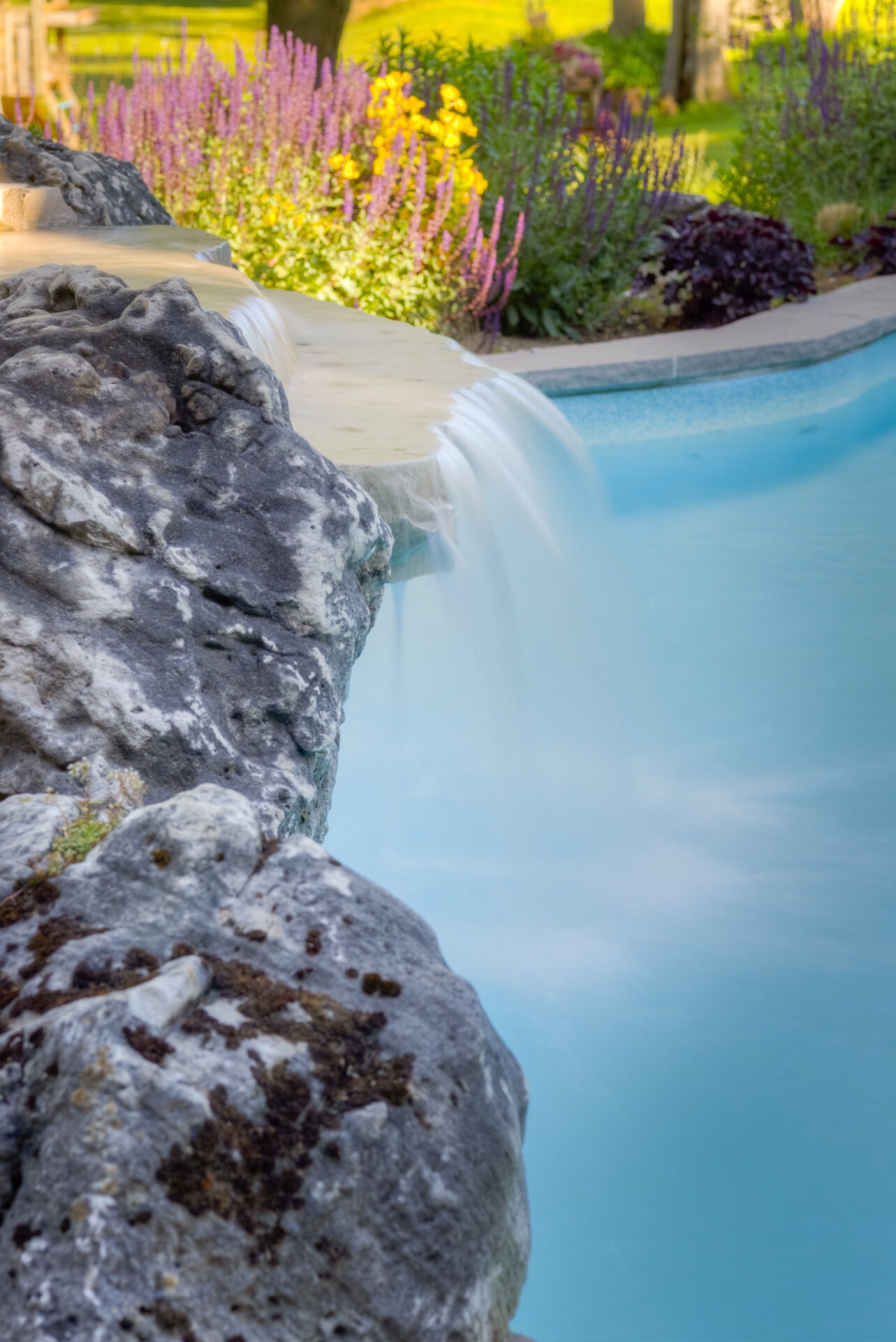 A tranquil waterfall cascades over rocks into a serene pool, surrounded by vibrant flowering plants and lush greenery in a landscaped garden.