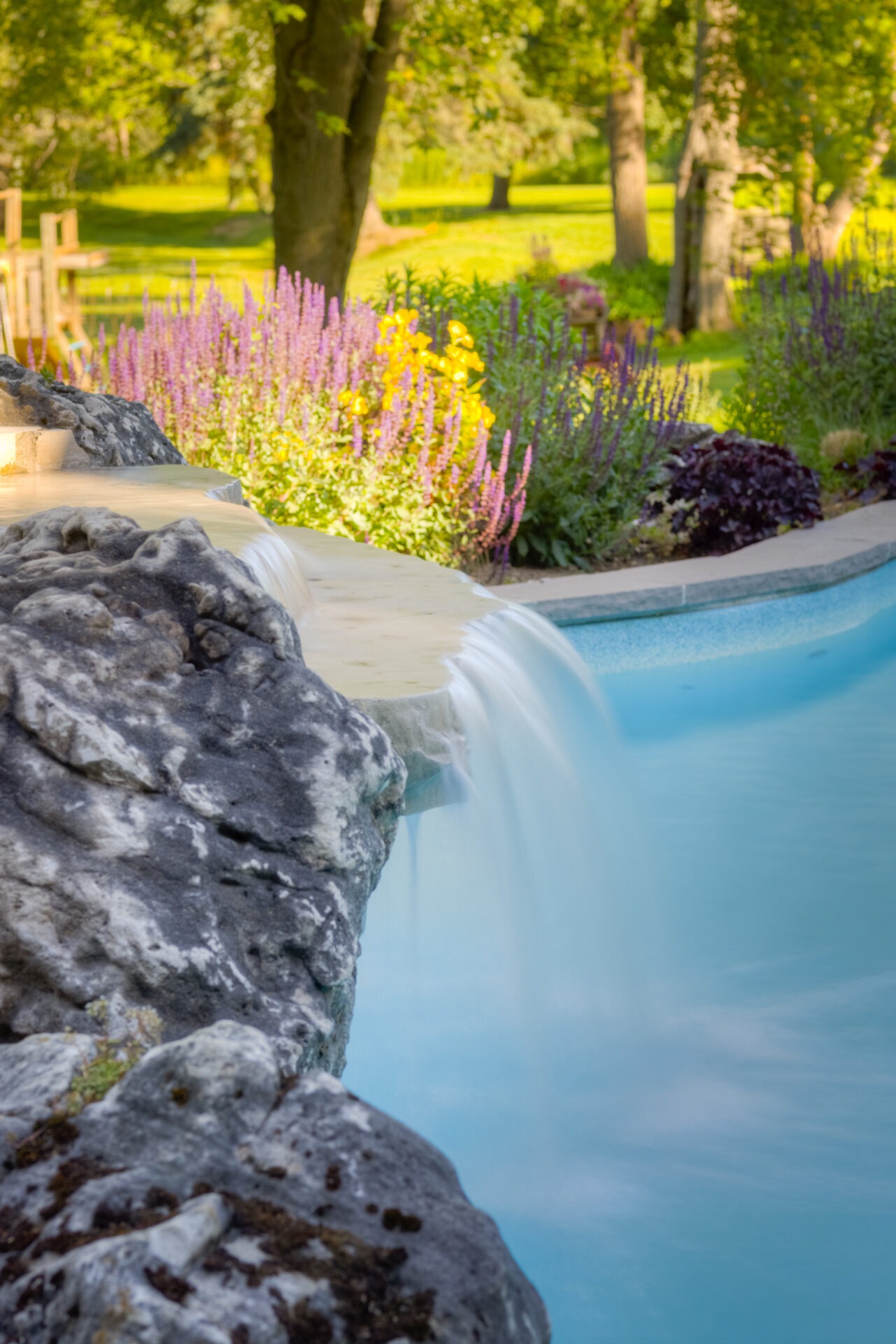 A serene artificial waterfall with clear blue water cascading over textured rocks, surrounded by lush colorful garden beds and green trees in the background.