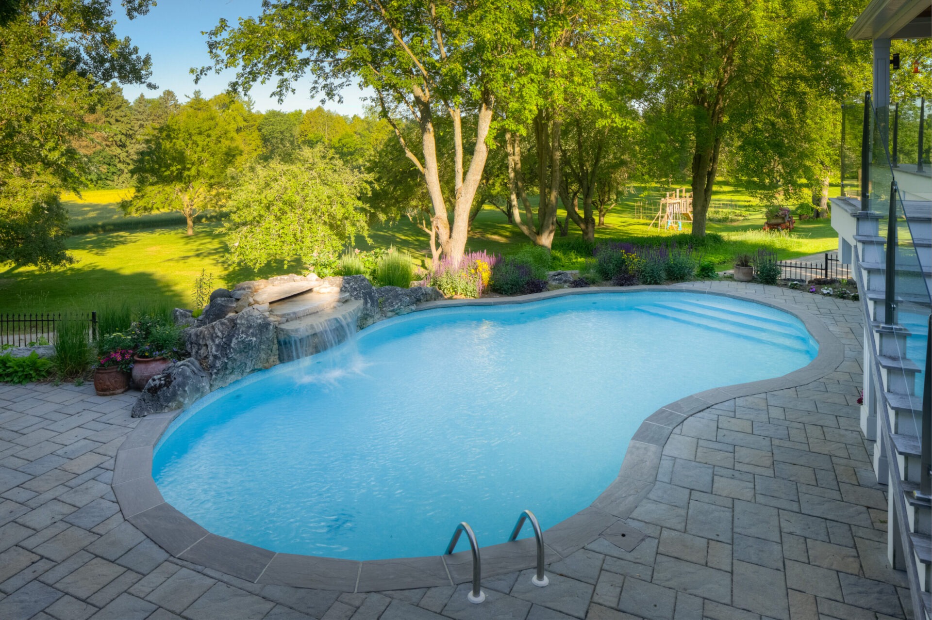An outdoor swimming pool with a waterfall feature, surrounded by a paver patio, lush greenery, and trees under a bright, sunny sky.