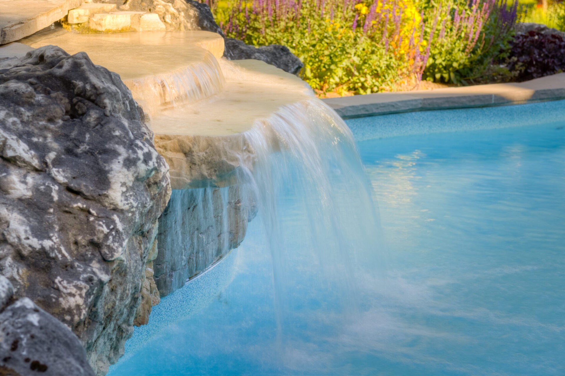 A serene waterfall flows from artificial stone into a clear, tranquil pool, surrounded by lush flowers in a landscaped garden at golden hour.
