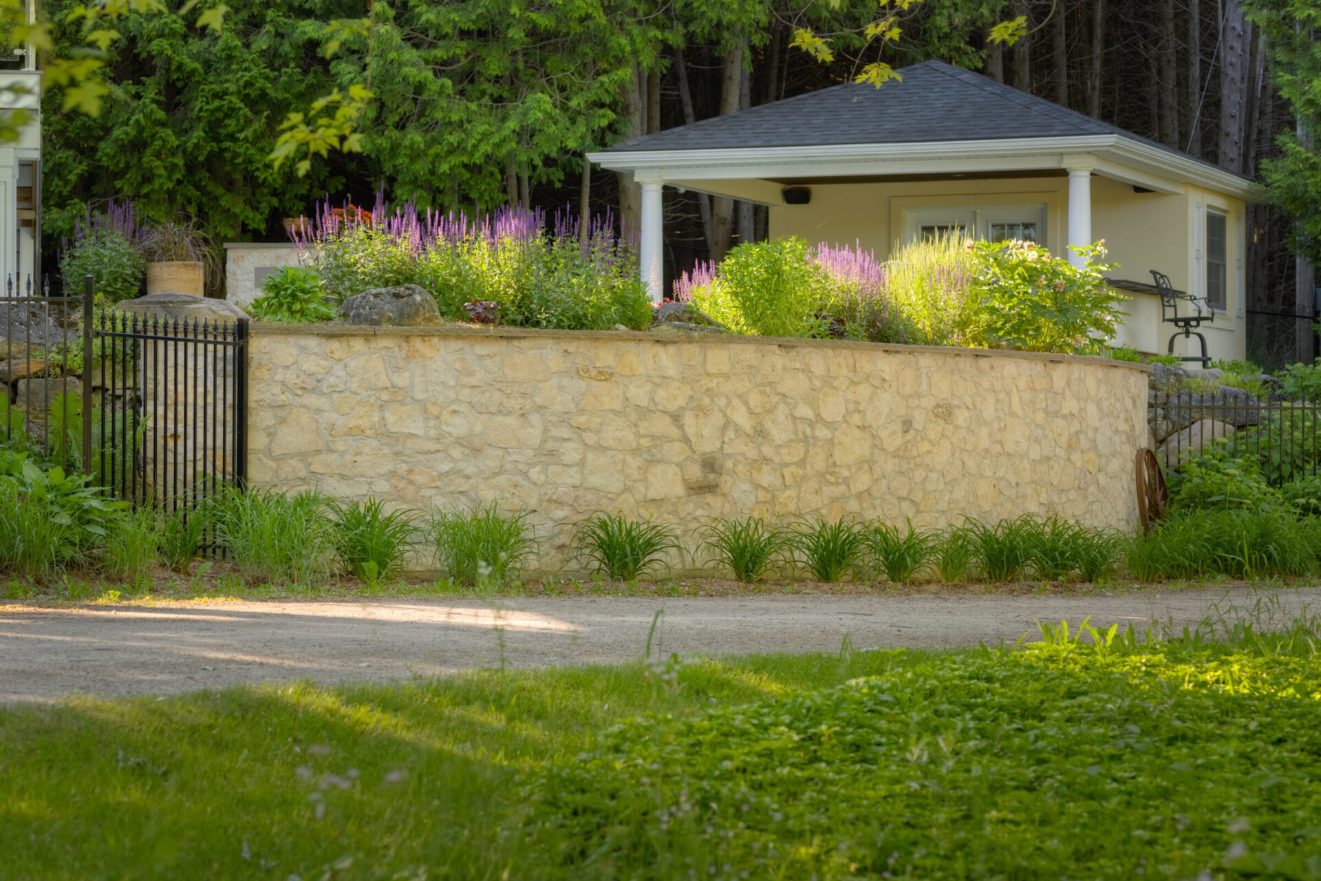 A stone garden wall adorned with purple flowers surrounds a residential property with a small building, lush greenery, and a metal gate.