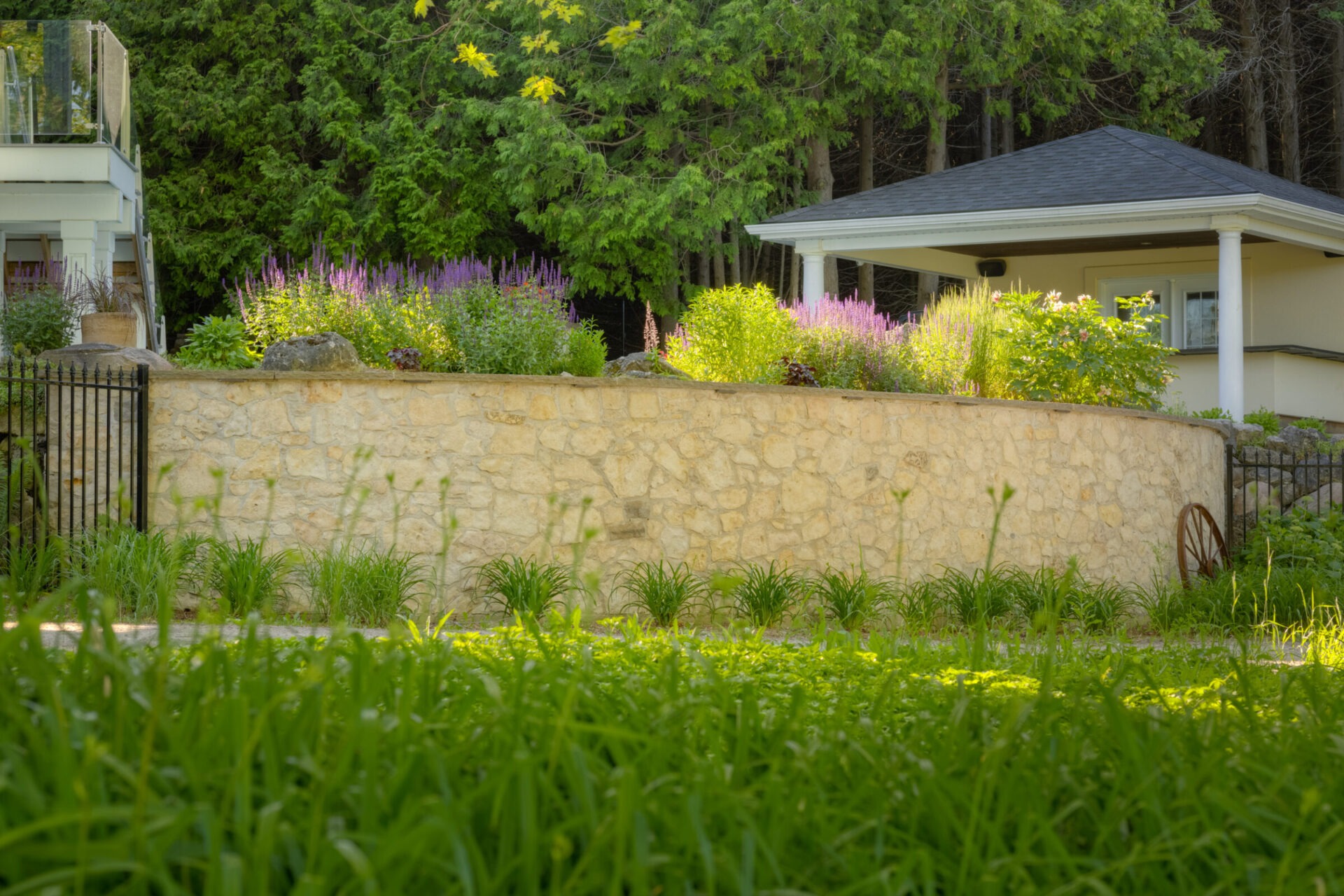 A stone retaining wall adorned with purple and green plants separates a lush lawn from a house partially obscured by trees and shrubbery.