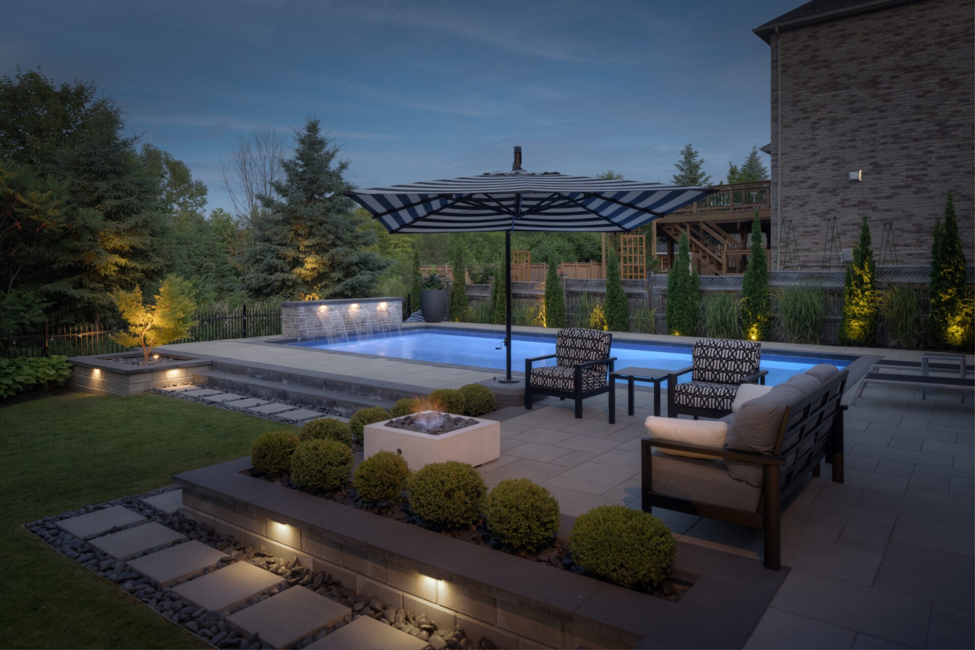 A tranquil evening setting featuring an illuminated swimming pool, elegantly patterned outdoor furniture, lush landscaping, and a stone fireplace in a residential backyard.