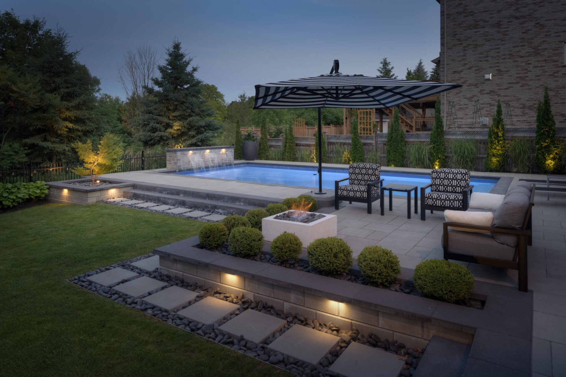 An elegant outdoor space at dusk featuring a swimming pool, patio with furniture, a fire pit, ambient lighting, and landscaped garden areas.