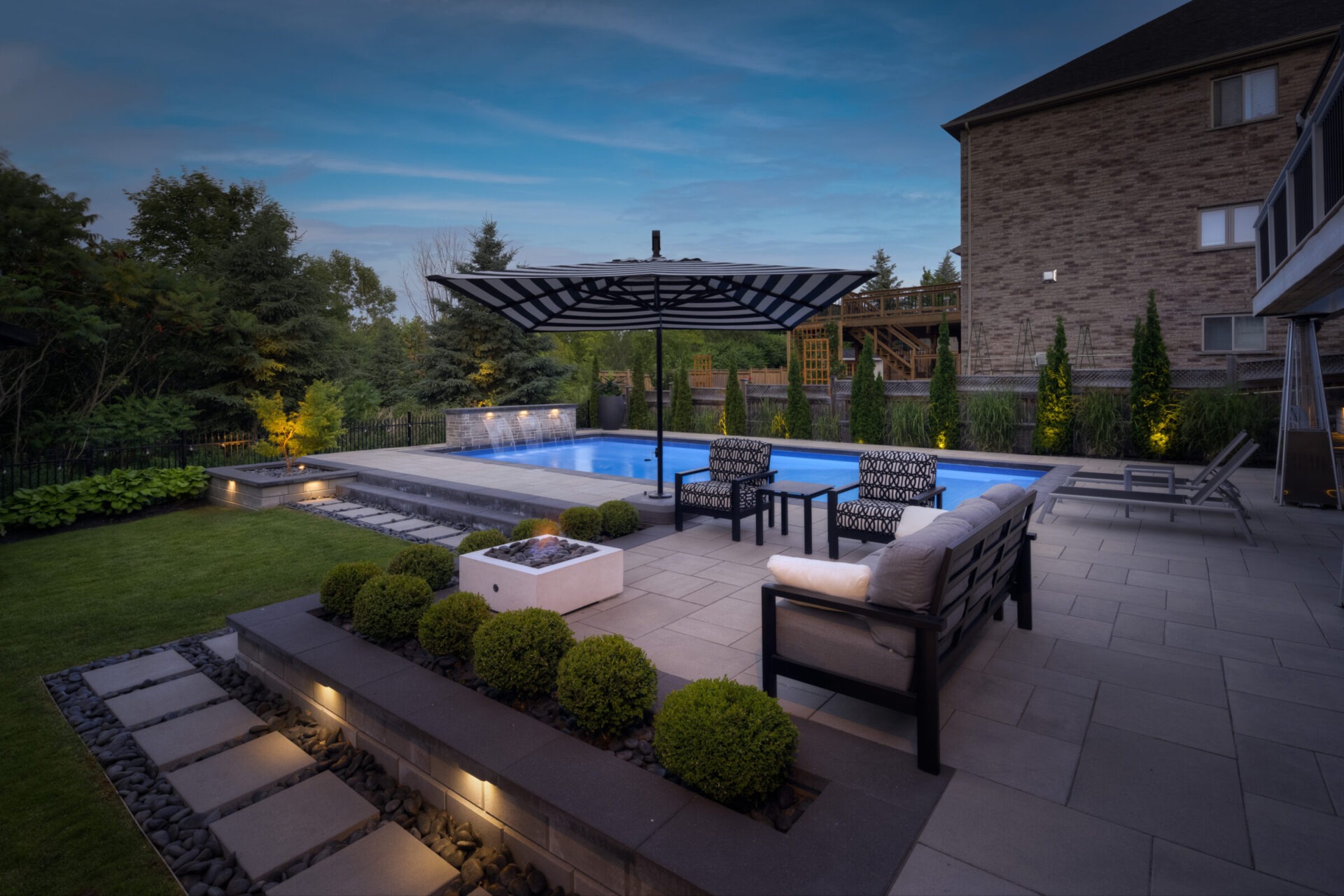 An elegant backyard at dusk featuring a swimming pool, patio with outdoor furniture, a pergola, manicured landscaping, and a brick house.