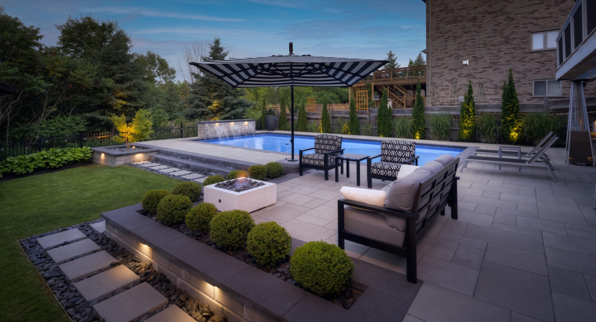 A serene backyard at dusk featuring a swimming pool, neatly landscaped garden, patio furniture, fire pit, and an elegant lounging area under an umbrella.